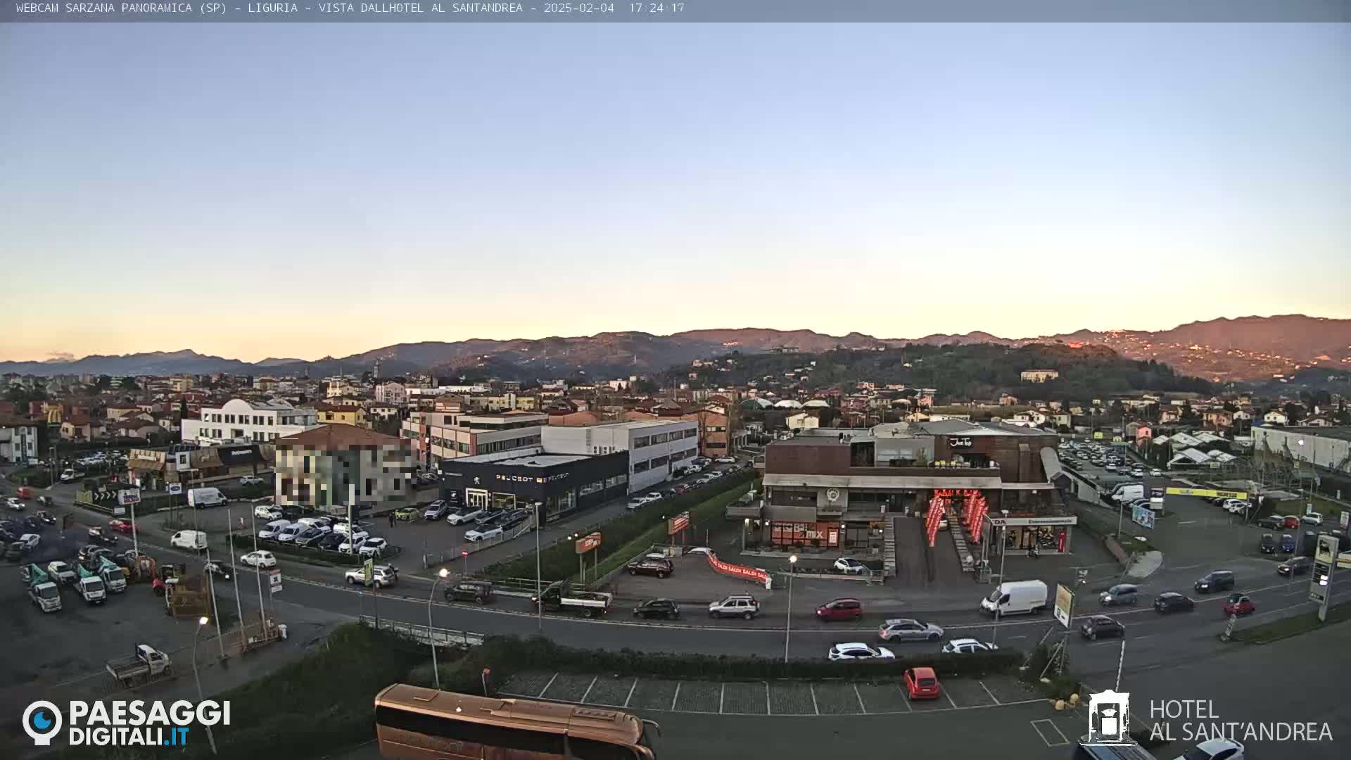 A panoramic view of a town at dusk, with mountains in the background and cars parked in a parking lot.