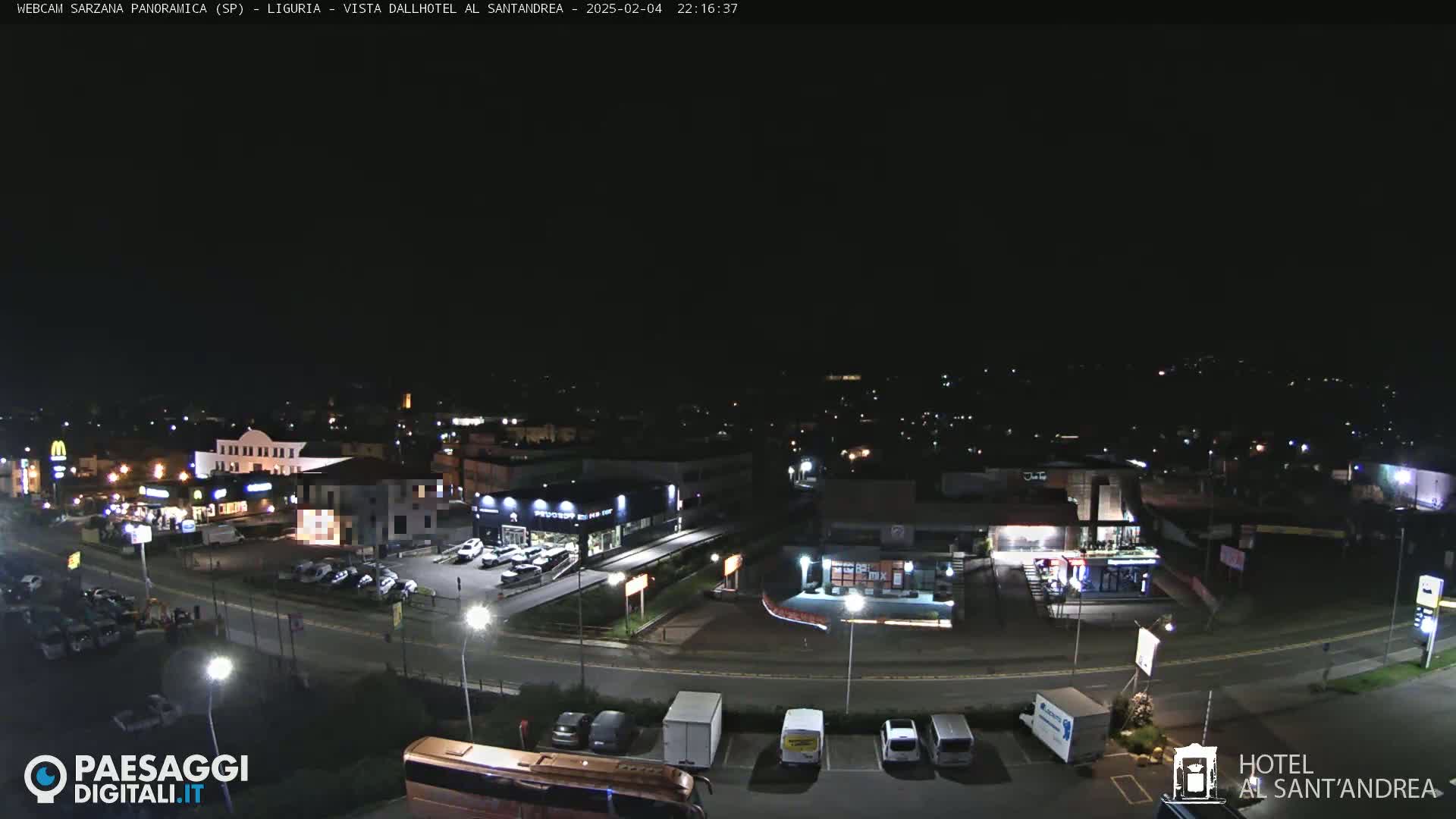 A nighttime, overhead view of a town's roads and businesses, including a parking lot with cars and a bus, under a clear, dark sky.