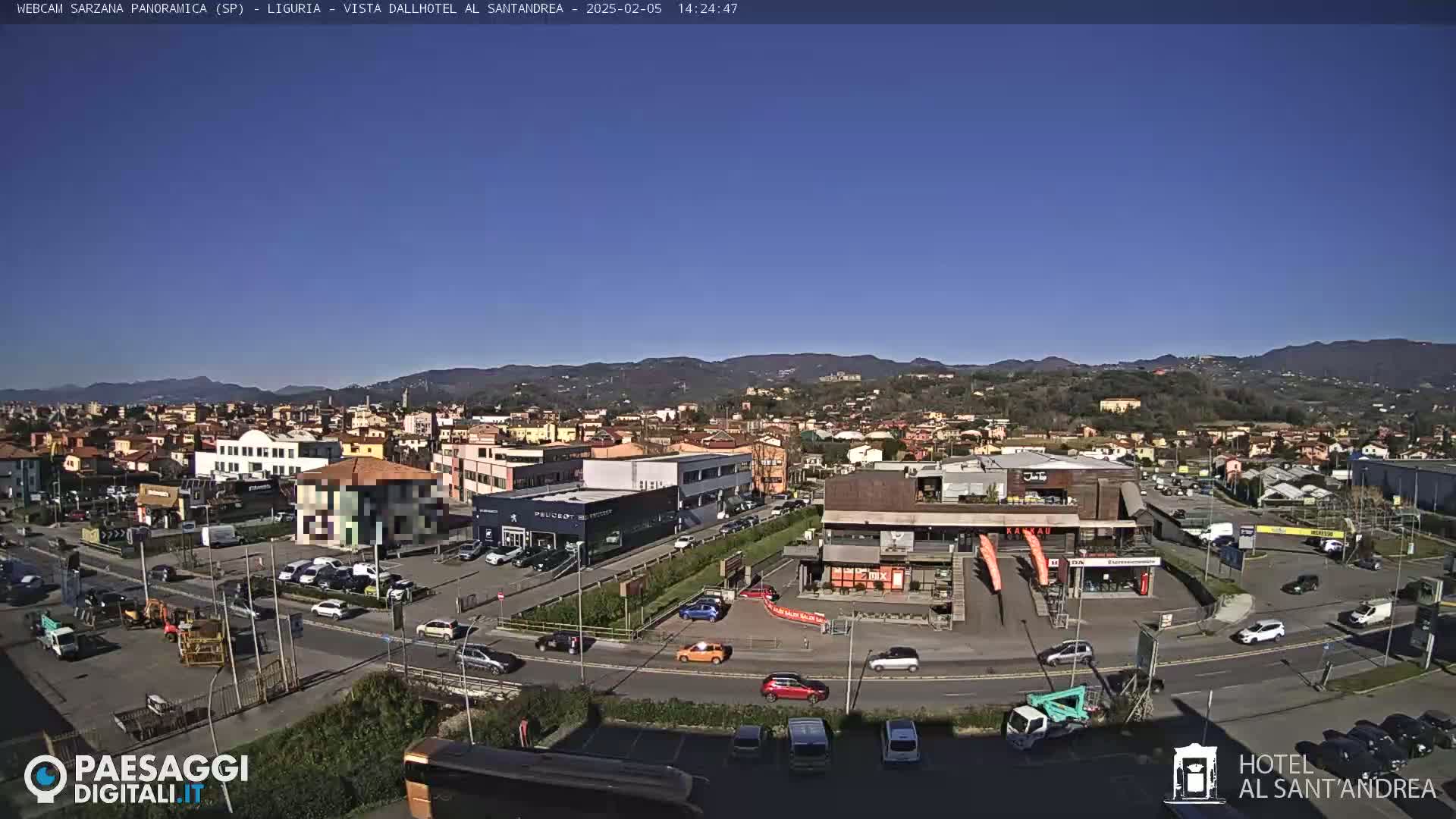 A sunny day reveals a panoramic view of a town with buildings, parking lots, and cars, nestled against a backdrop of hills.