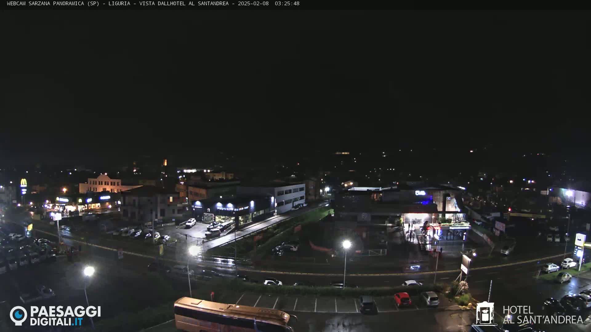 A nighttime, rain-slicked view of a town's parking lots and businesses, with several cars visible.