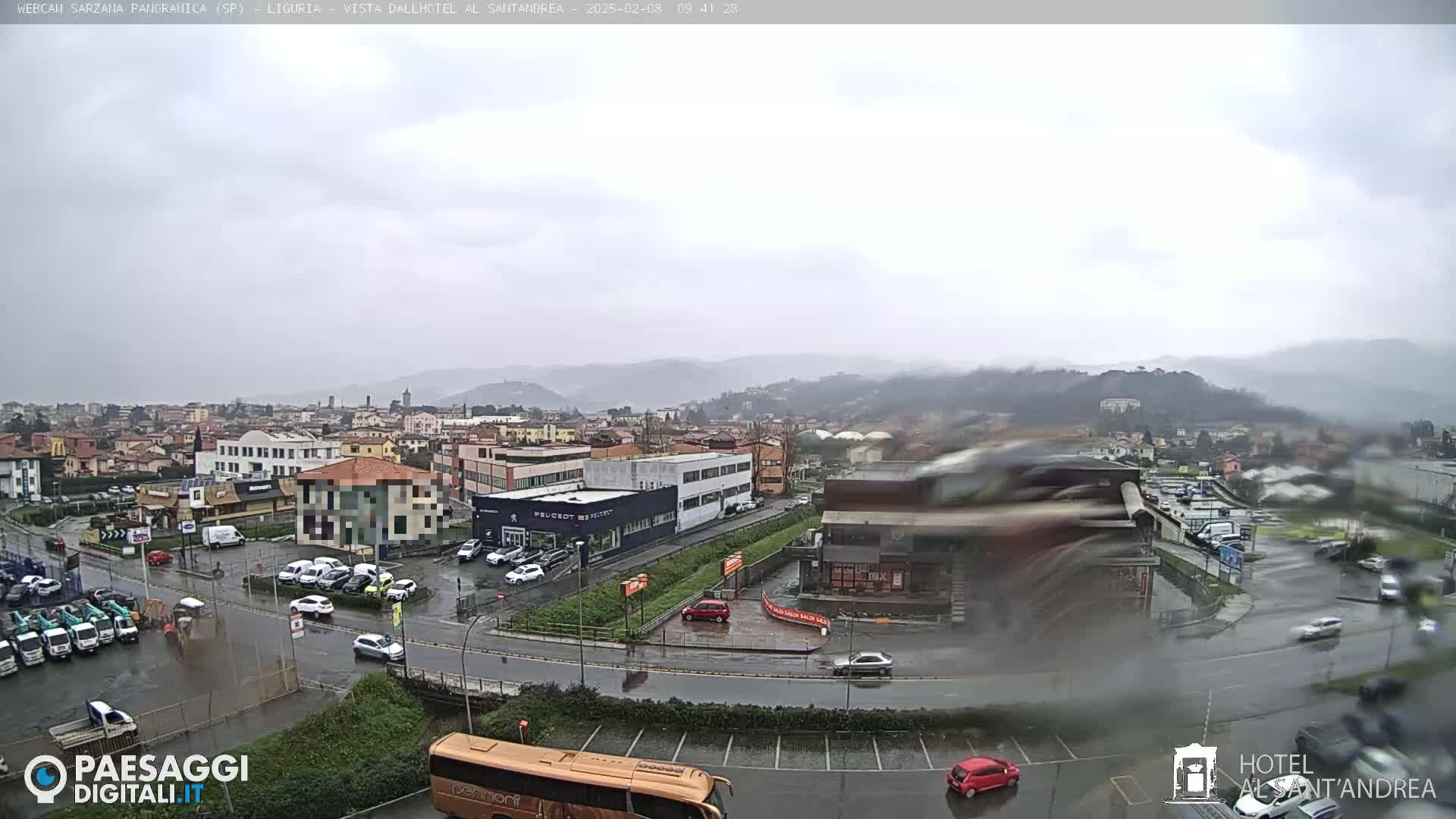 A rain-soaked town sprawls beneath a gray sky, with cars parked in lots and driving on wet roads.