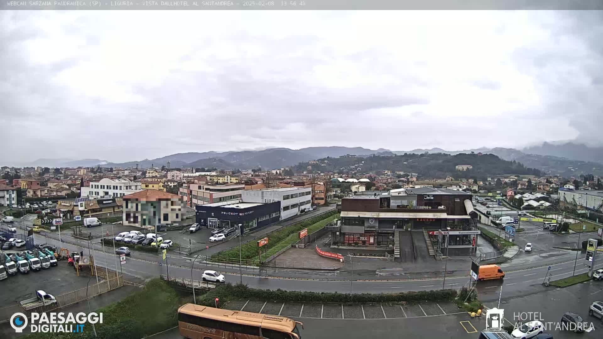 A wide, high-angle view of a town on a cloudy day, showing numerous buildings, parking lots filled with cars, and a bus, with mountains visible in the background.