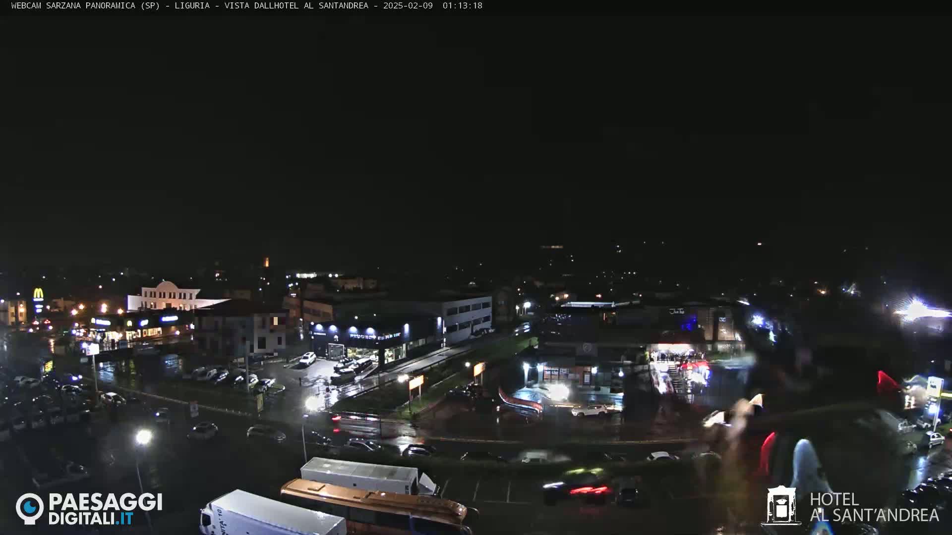 A nighttime aerial view of a town shows wet roads and illuminated buildings under a dark sky.