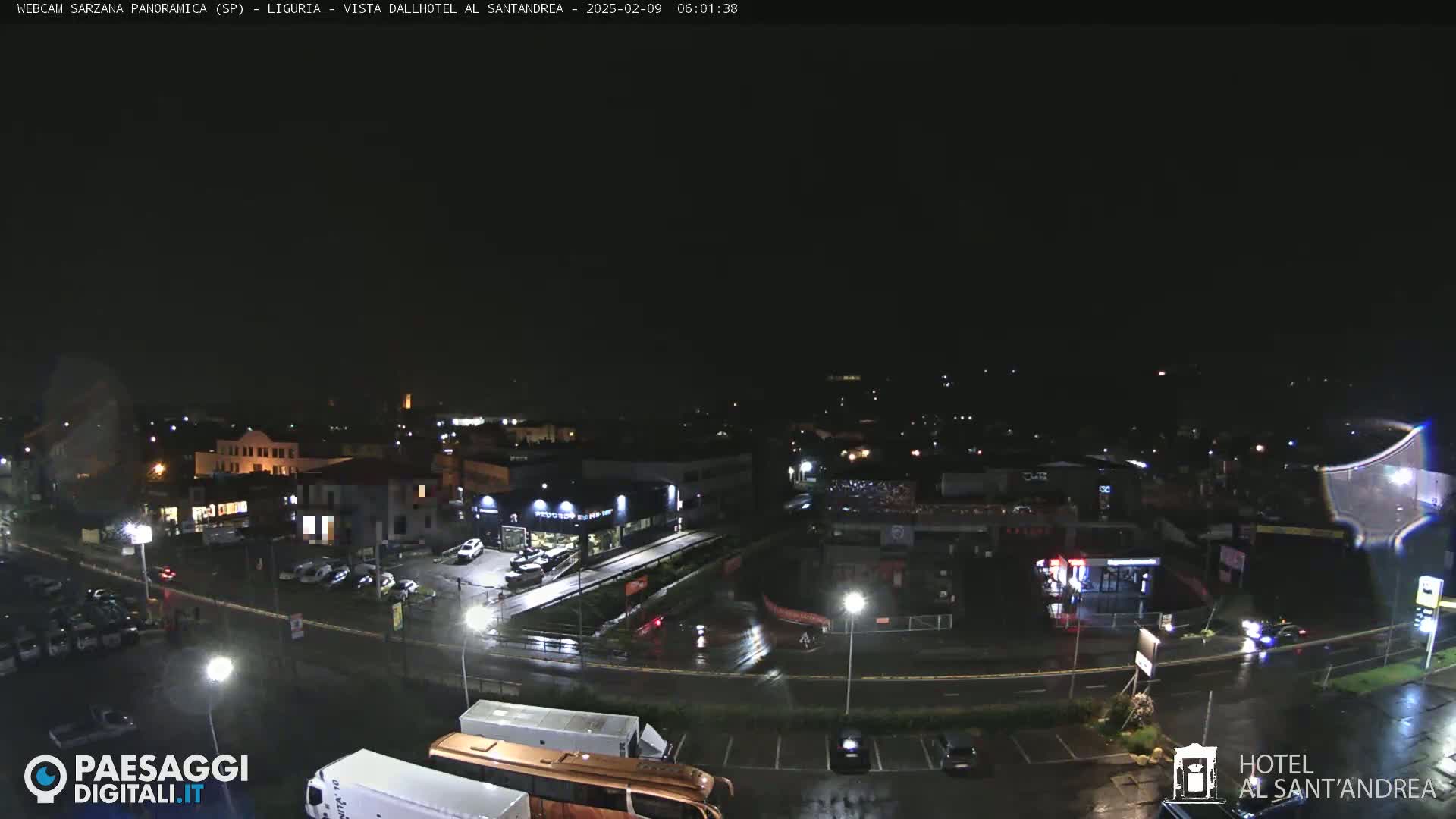 A nighttime, rain-slicked view of a town's streets and buildings, with cars parked in lots and driving on roads.