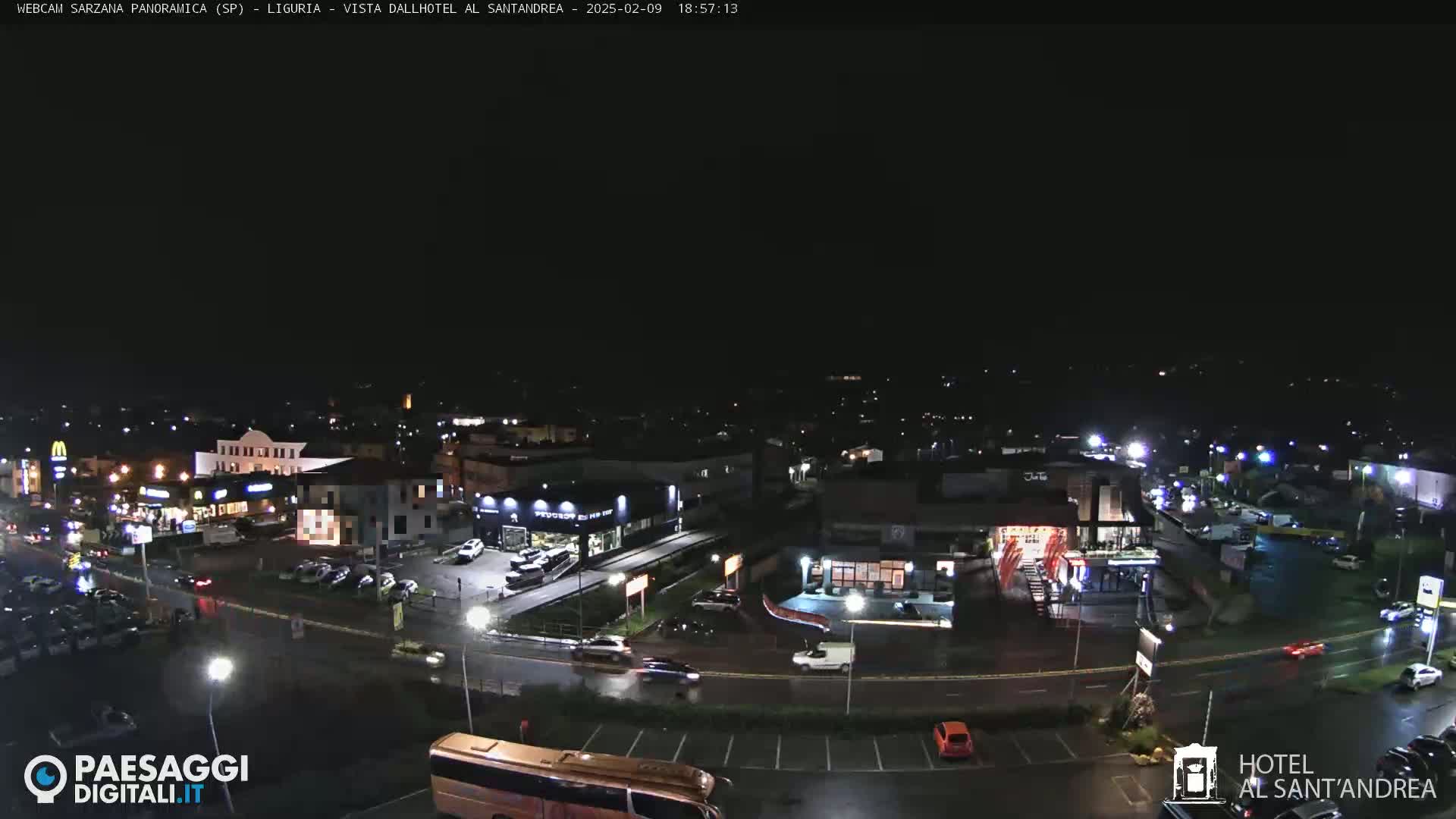 A nighttime, wet aerial view of a town with illuminated buildings and vehicles, showing a road intersection and parking lots.