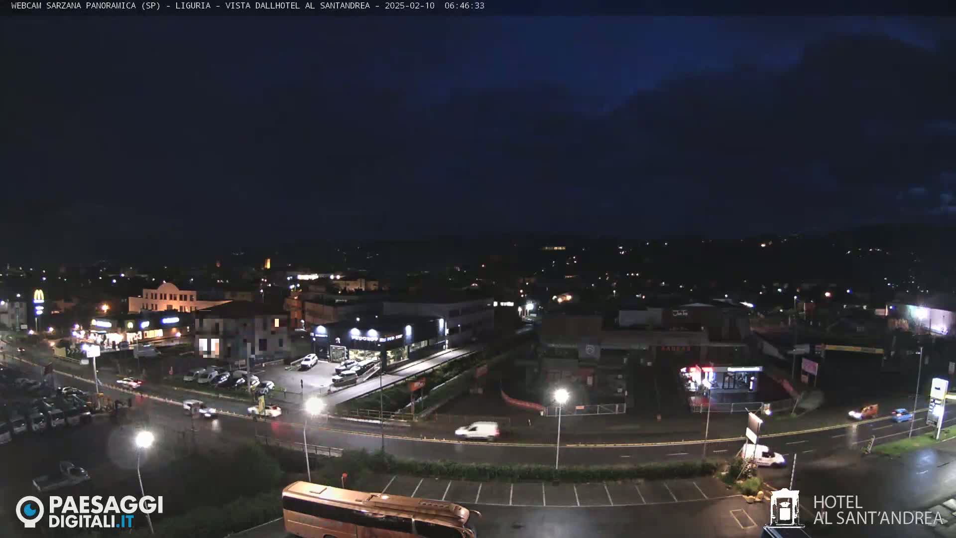 A nighttime, slightly rainy view of a town with illuminated buildings and roads, featuring cars and a bus in a parking lot.