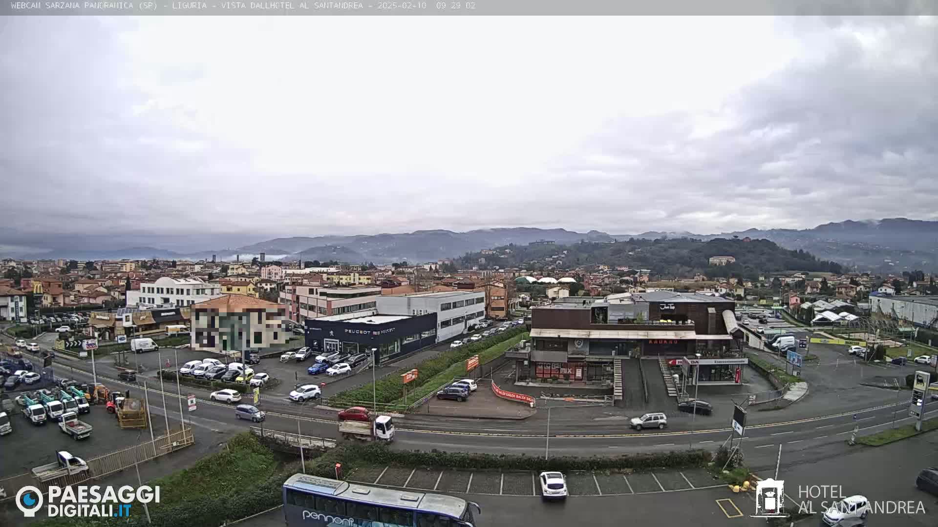 A wide, aerial view of a town under an overcast sky, showing a parking lot with cars, buildings, and a bus, with hills and mountains in the background.