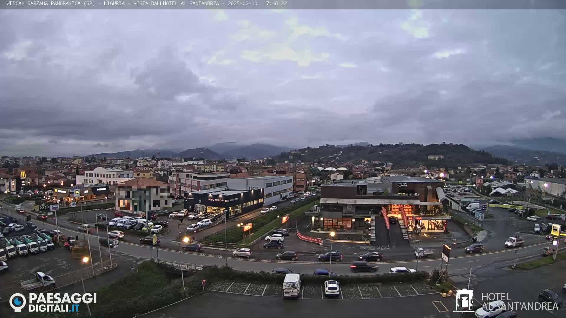 A mostly cloudy, overcast day reveals a town's cityscape with several buildings, cars, and parking lots, situated before a range of hills.