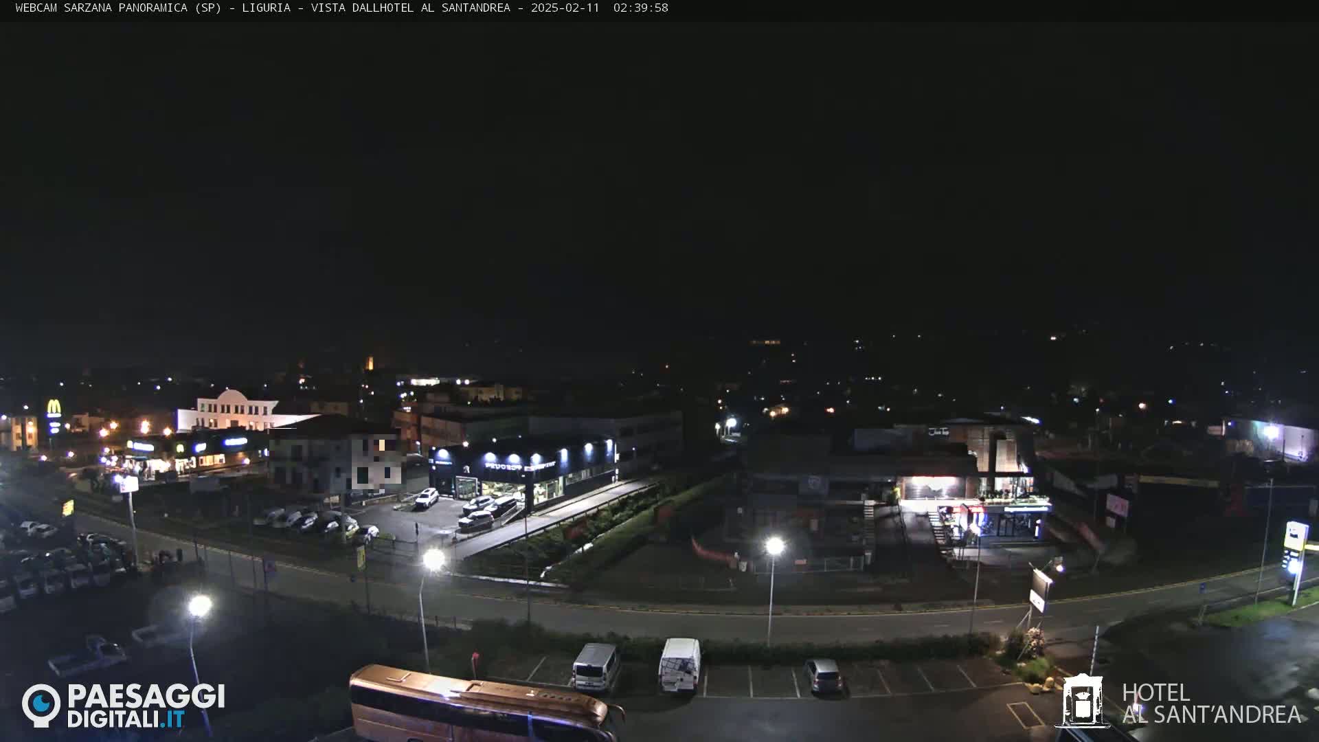A nighttime, wide shot of a town's outskirts, showing a road curving through parking lots and businesses with some cars visible.