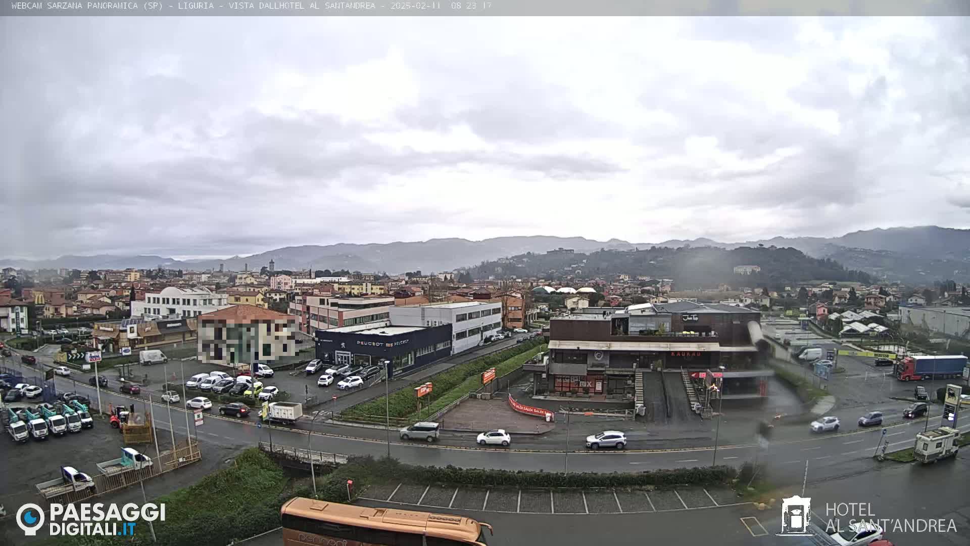A cloudy day reveals a panoramic view of a town, featuring numerous buildings, vehicles in parking lots and on roads, and distant mountains under a gray sky.