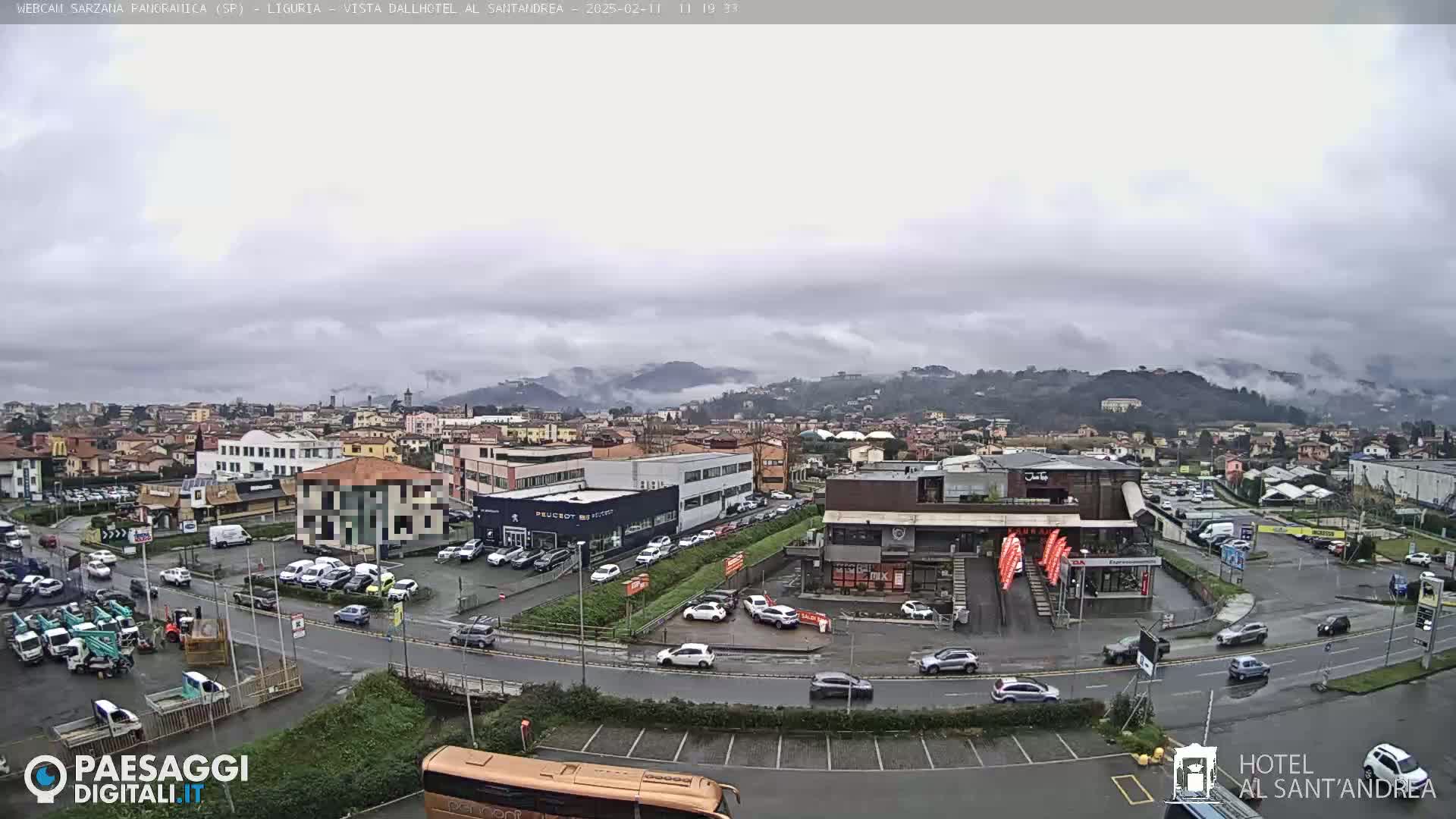 A cloudy, overcast day reveals a panoramic view of a town with numerous buildings, parking lots filled with cars, and a road network, all nestled against a backdrop of rolling hills partially shrouded in fog.