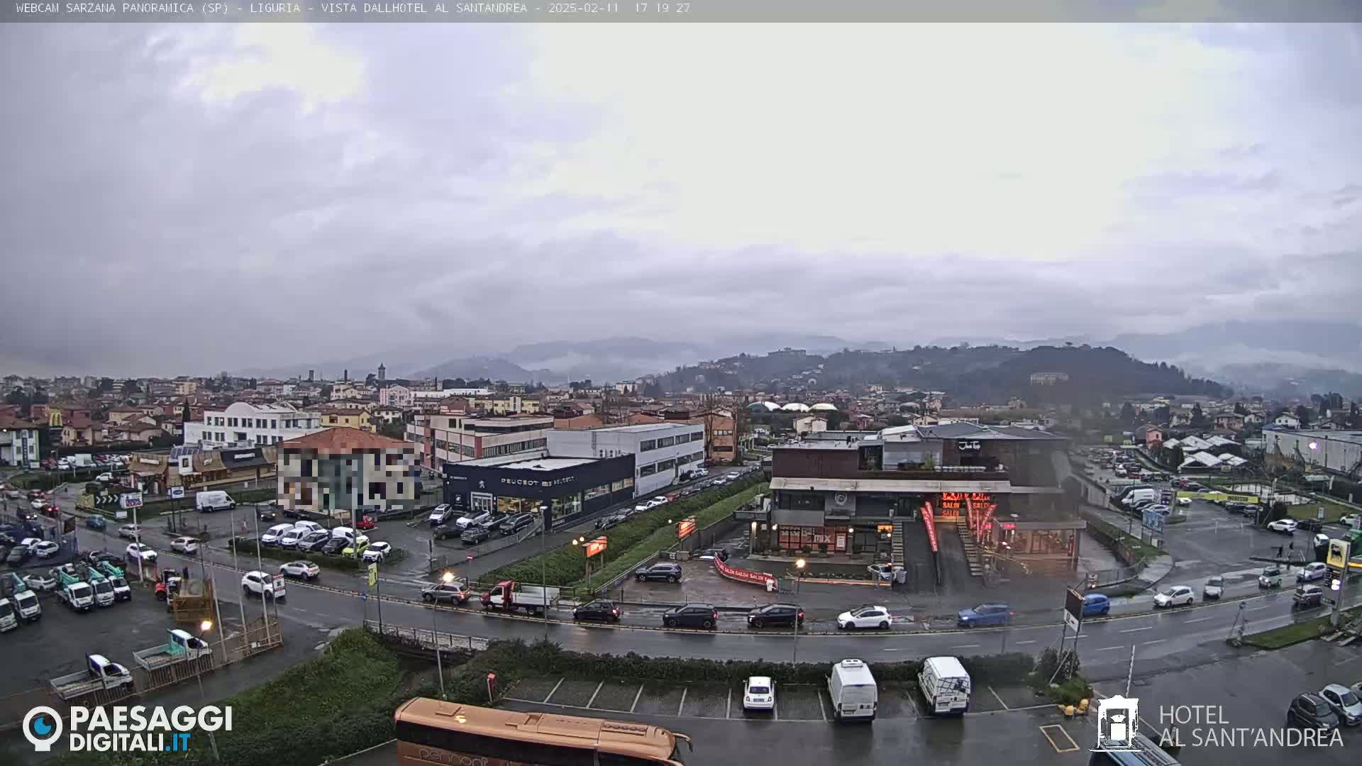 A wide, overhead view of a town under an overcast sky shows many cars parked in lots and driving on wet roads.