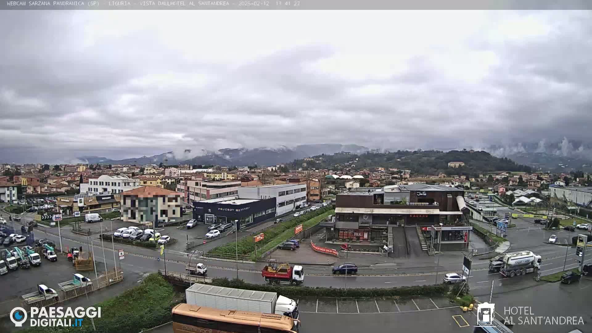A wide shot of a town on an overcast day, showing a mix of buildings, vehicles, and parking lots, with hills and mountains visible in the background.