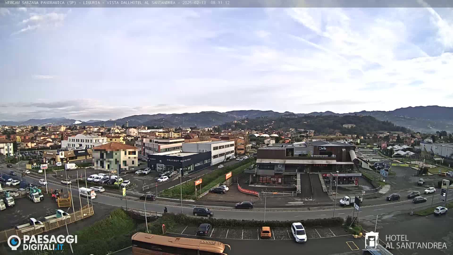 A panoramic view of a town on a partly cloudy day, showing buildings, roads with cars, and distant mountains.