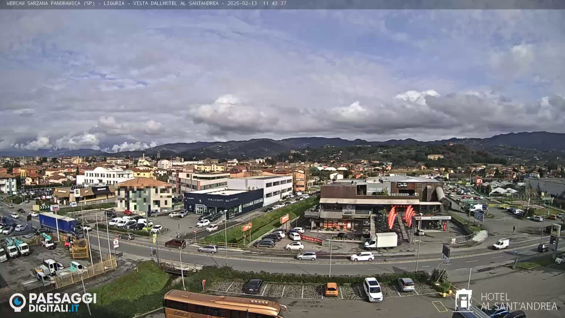 A partly cloudy day reveals a panoramic view of a town nestled amongst hills, with a roadway curving through a parking lot filled with cars and commercial buildings.