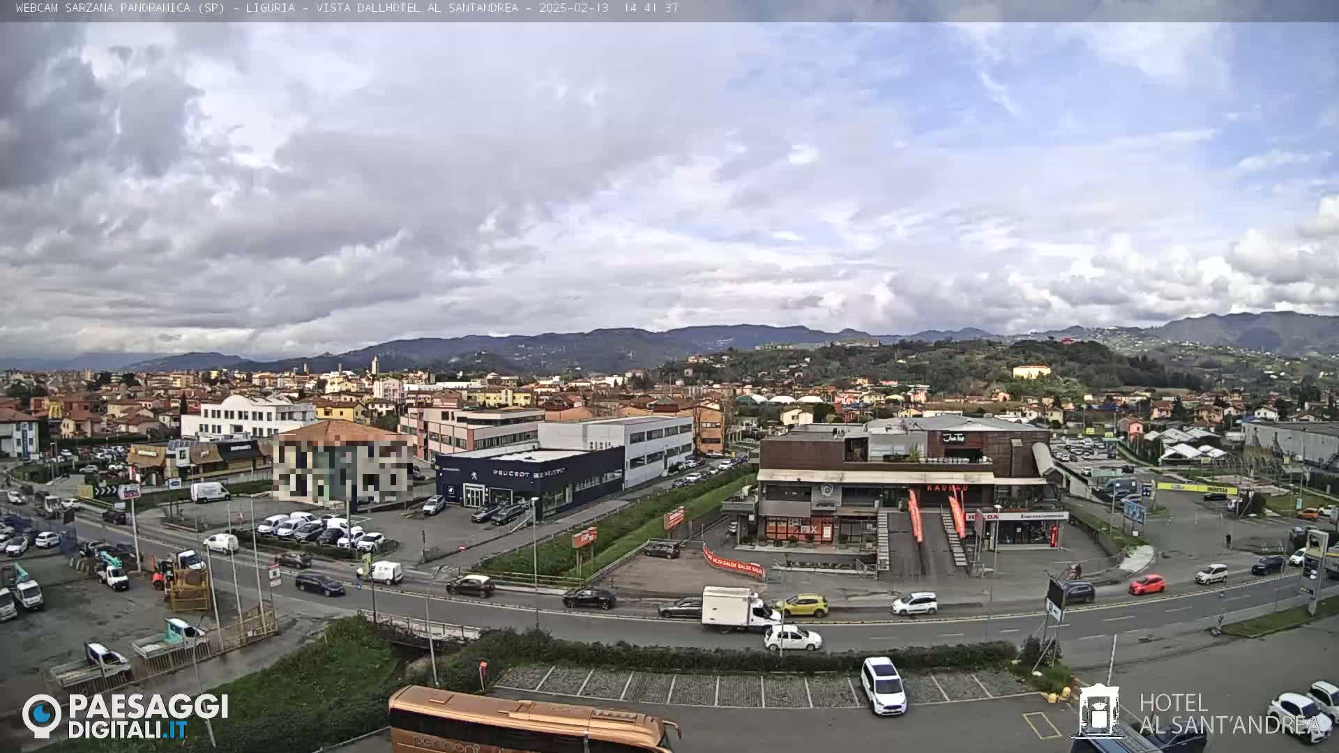 A partly cloudy day reveals a panoramic view of a town nestled among hills, with cars parked in lots and driving on roads near a variety of buildings.