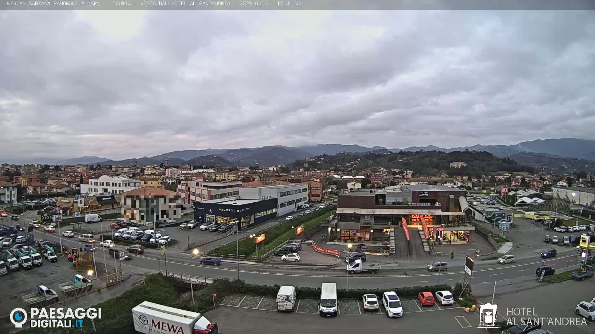 A mostly cloudy day reveals a view of a town nestled in a valley, surrounded by hills and mountains, with cars parked in a lot near several buildings.