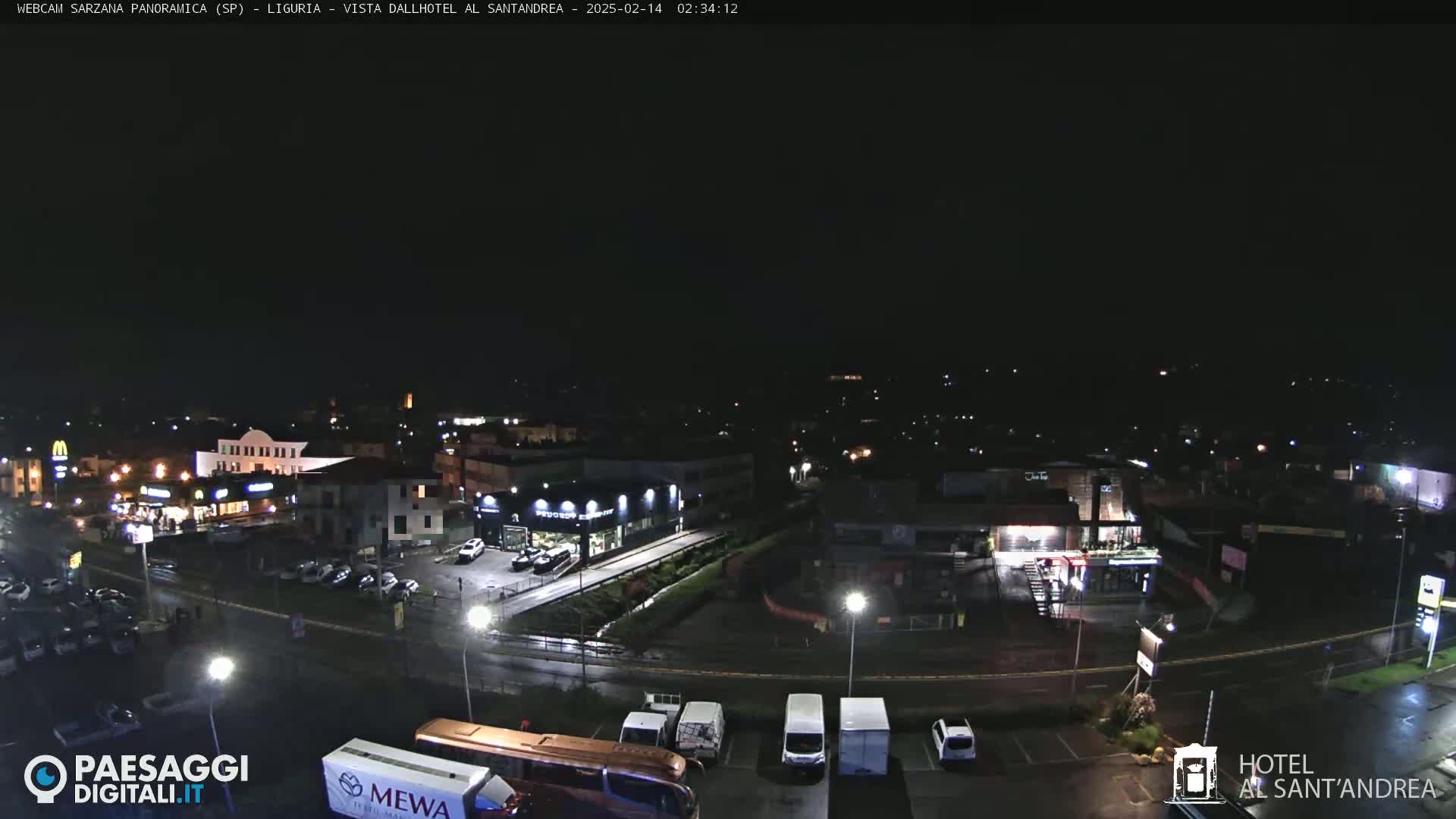 A nighttime view of a town with wet roads, showing buildings, parked cars, and a large bus.