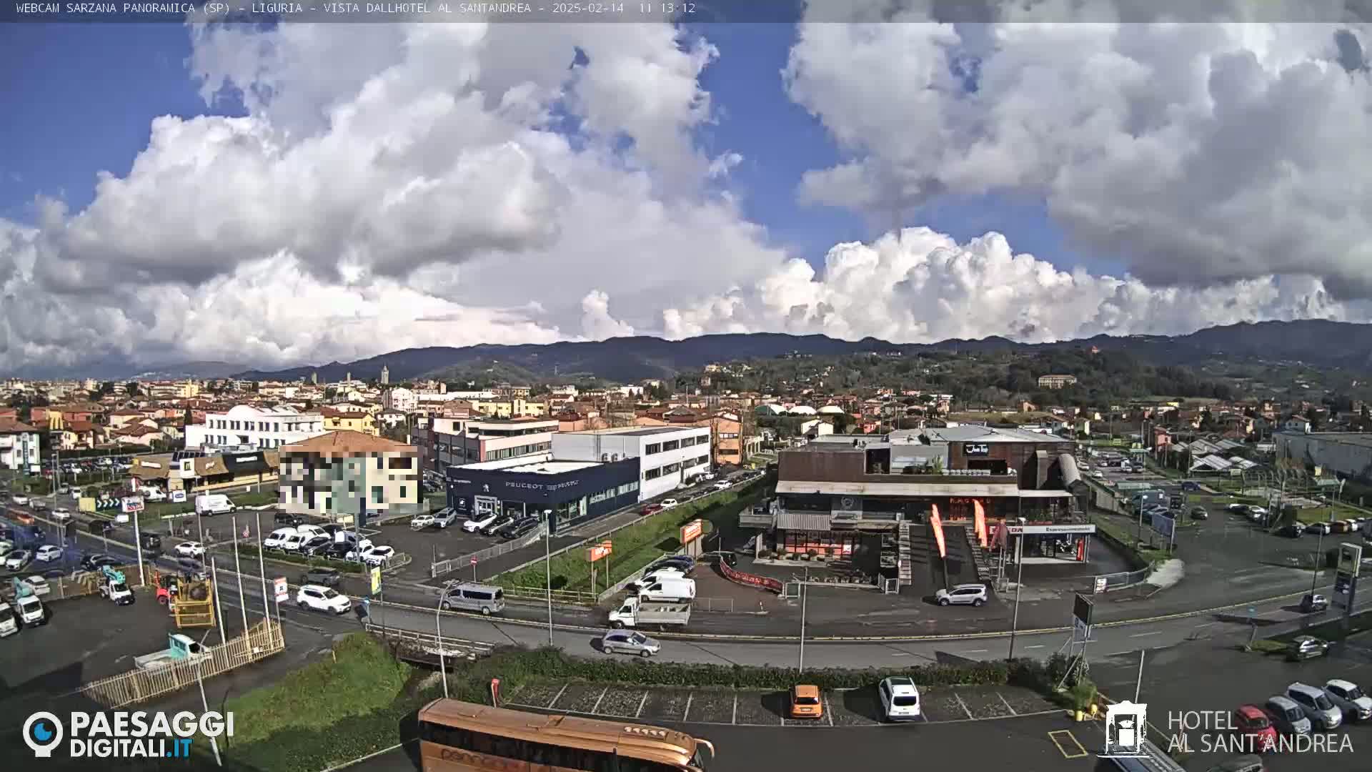 A partly cloudy day reveals a panoramic view of a town nestled between hills, with traffic visible on roads and in parking lots.