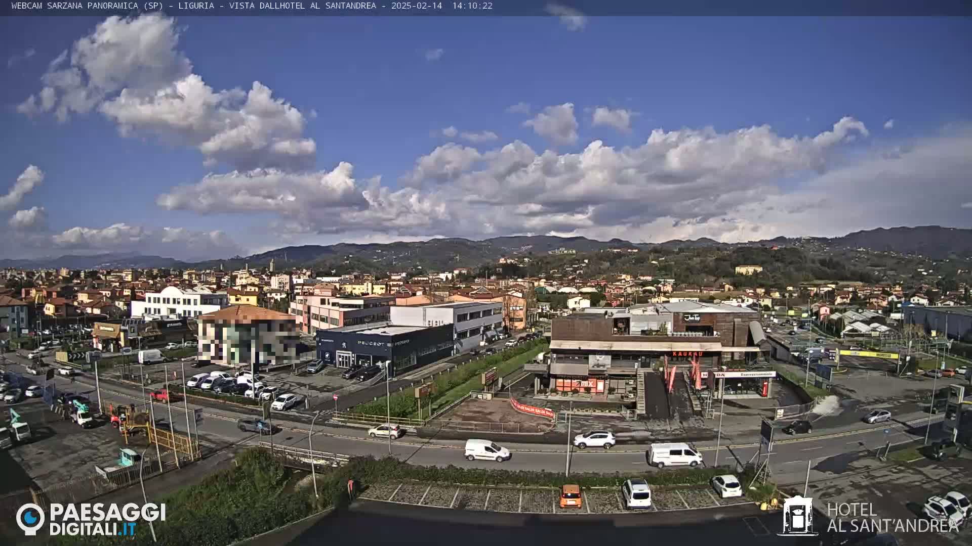 A partly cloudy day reveals a view of a town nestled between hills, with cars parked in a lot and moving along roads.