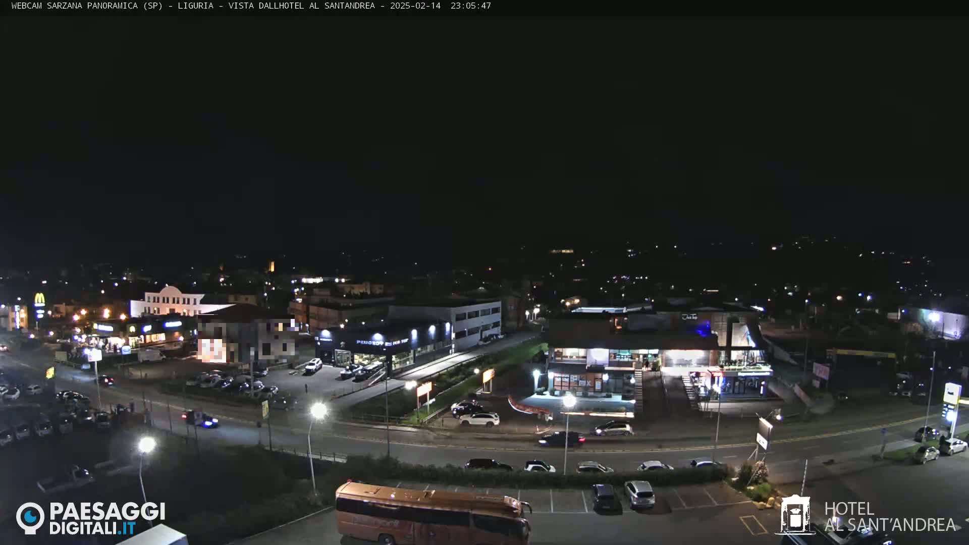 A nighttime aerial view of a town, showing roads, buildings, parked cars, and a large orange bus, under a clear, dark sky.