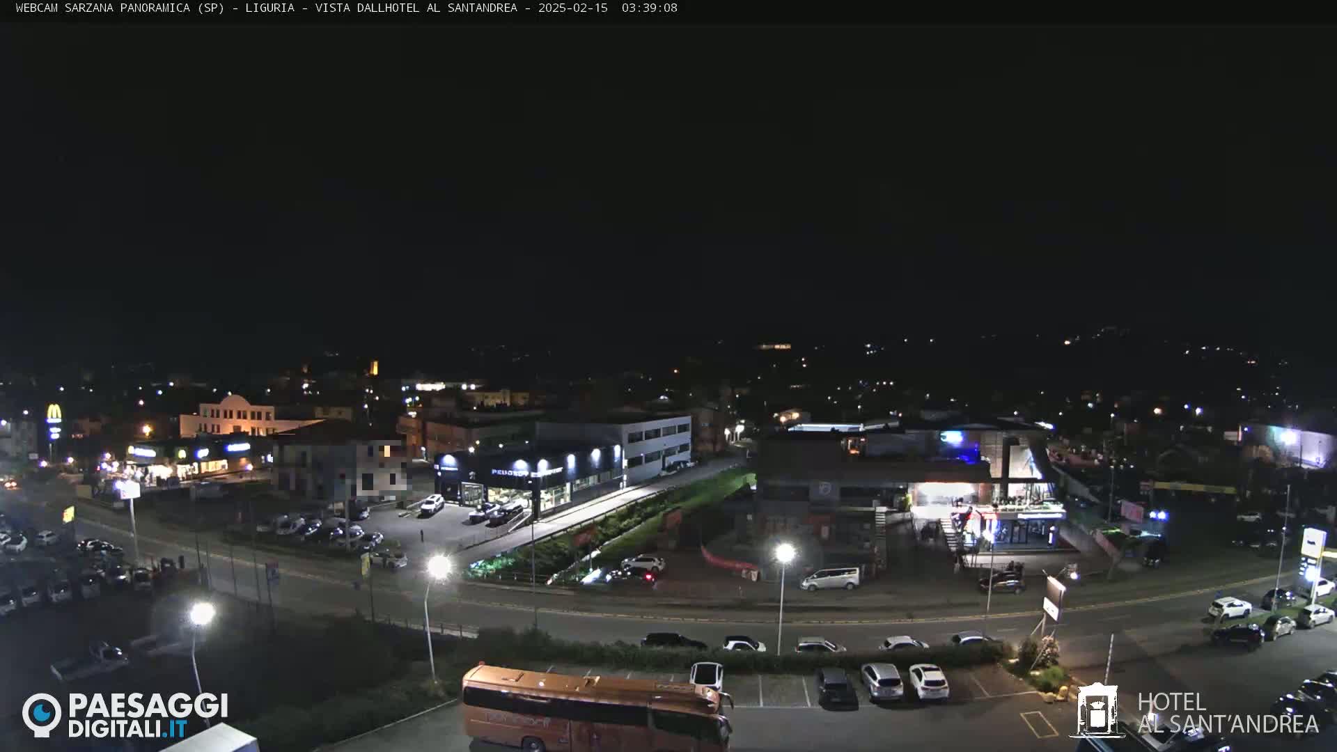 A nighttime aerial view of a town shows roads, buildings, parking lots, and vehicles under a clear, dark sky.