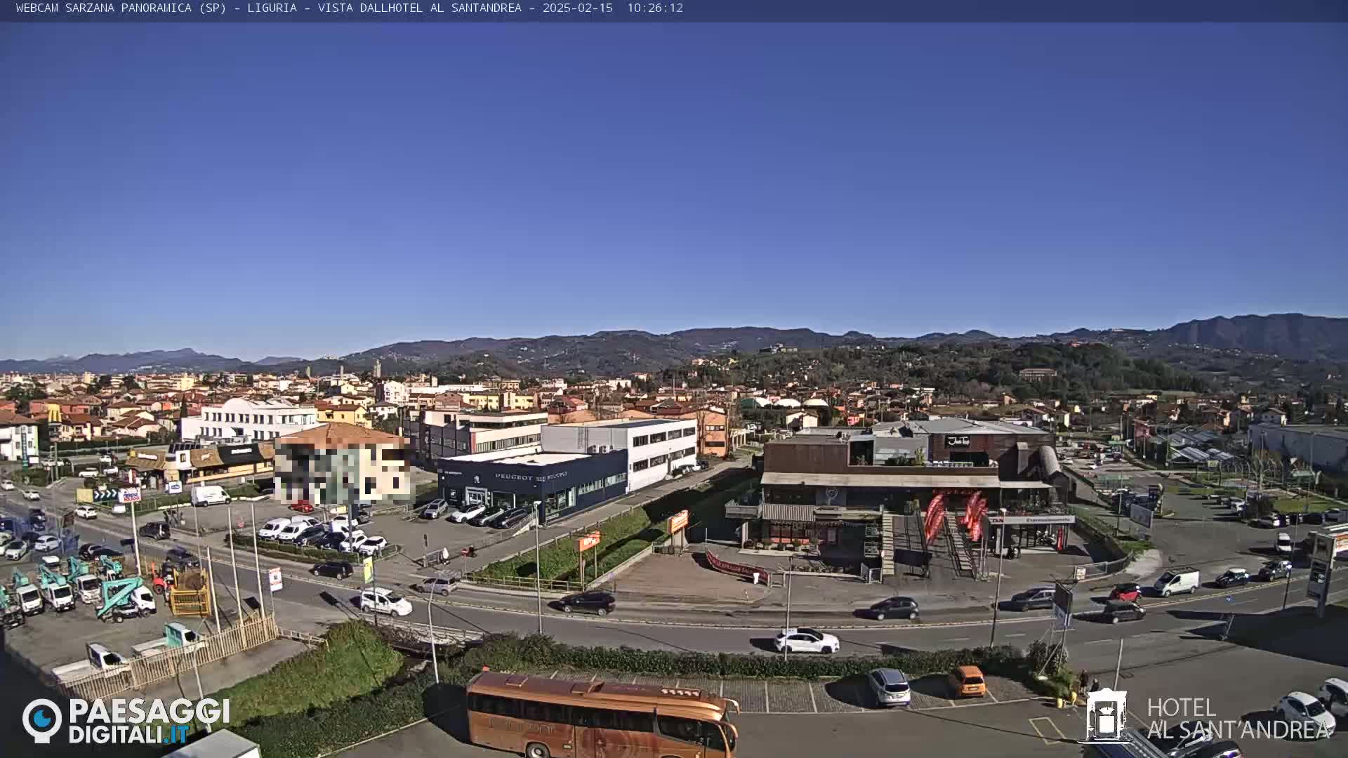 A sunny day reveals a panoramic view of a town nestled among hills, with cars parked in lots and driving on roads surrounding various buildings, including what appears to be a hotel and car dealership.