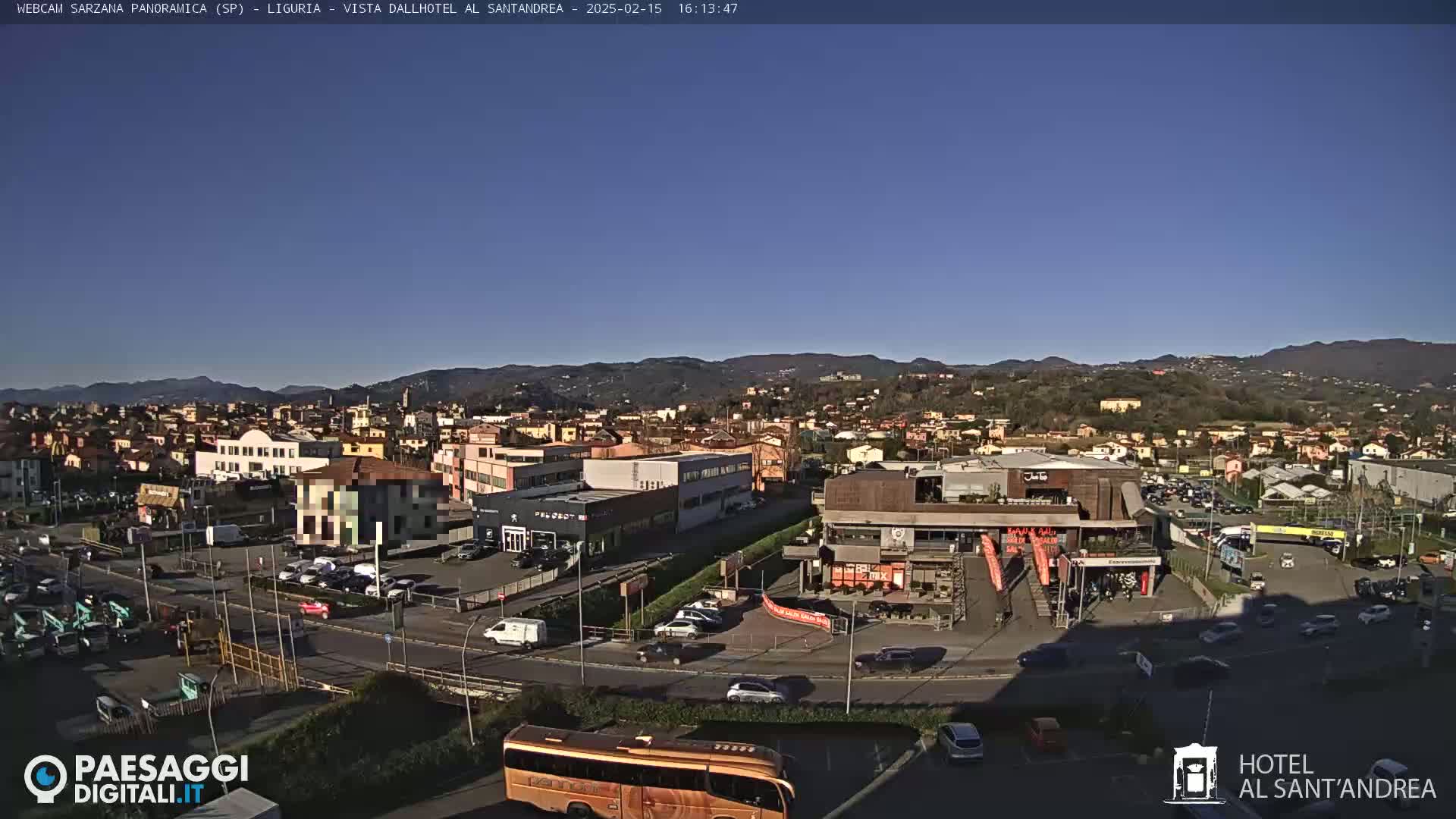A sunny, clear day reveals a wide shot of a town nestled in a valley, with mountains in the distance, featuring buildings, parked cars, and a road with several vehicles.