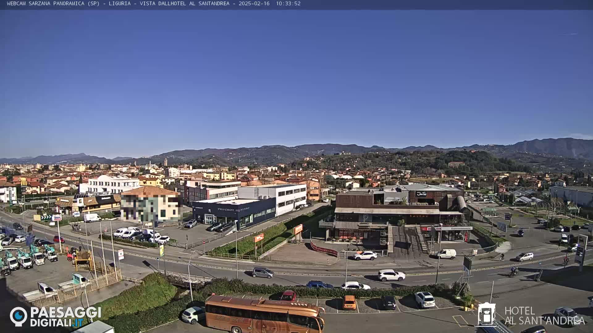 A sunny day reveals a sprawling town nestled against a backdrop of distant mountains, with cars parked in lots surrounding various buildings and a large bus visible in the foreground.