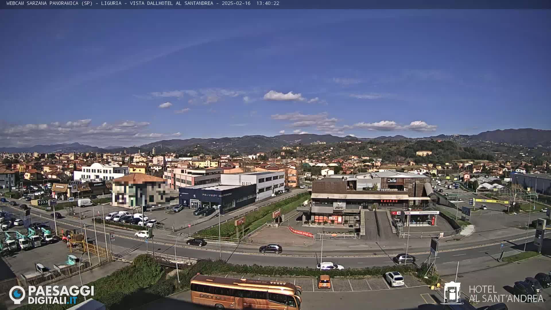 A sunny day reveals a sprawling town nestled in a valley, surrounded by mountains, with cars parked in lots near buildings and a large bus visible in the foreground.