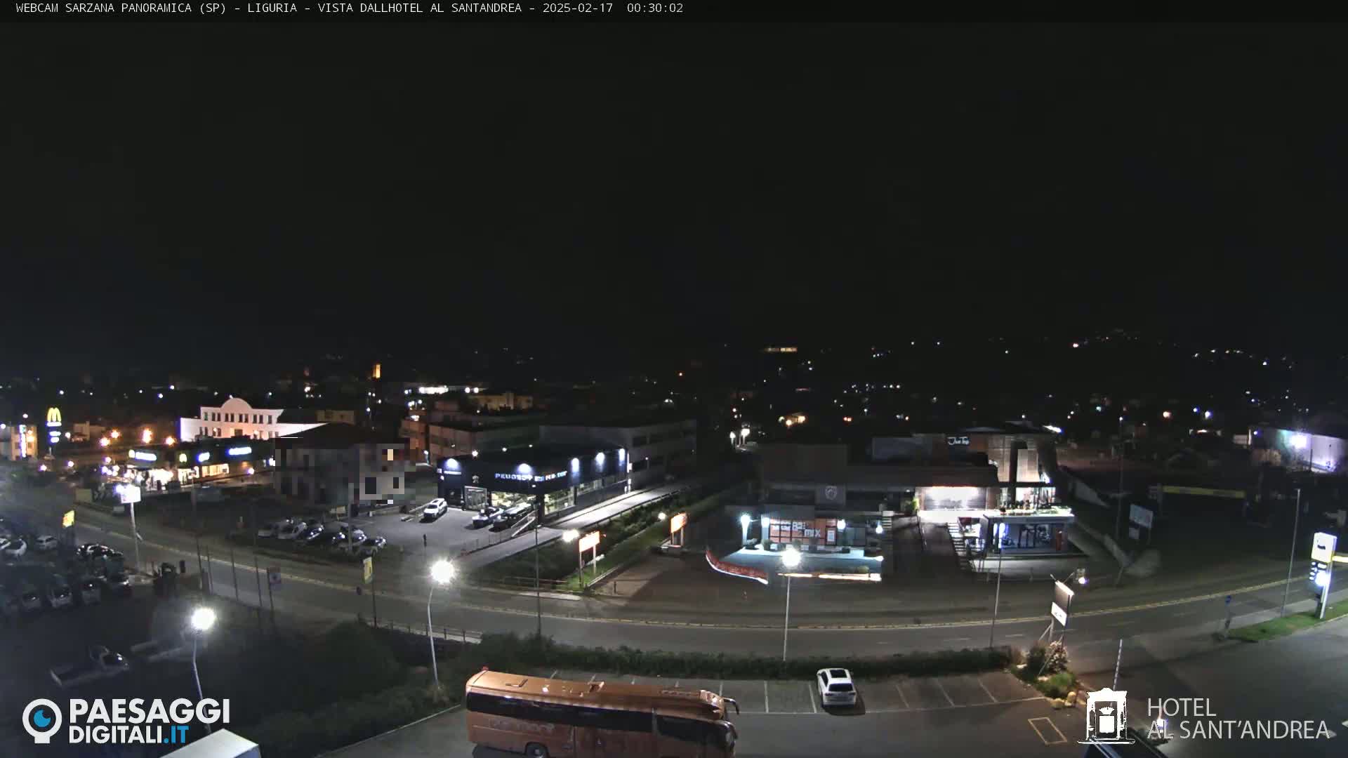 A nighttime, high-angle view of a town's outskirts, showing a roadway curving through a parking lot with several cars and a bus, and various businesses and buildings with lights visible.