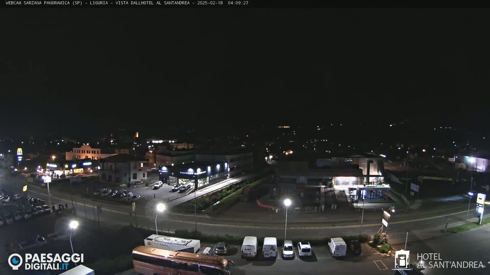A nighttime, overhead view of a town's streets and businesses, with cars parked in lots and a bus in the foreground.