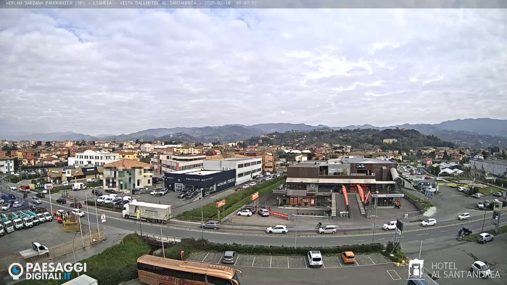 A mostly cloudy day reveals a panoramic view of a town, featuring buildings, roads, and parked cars, with mountains visible in the background.