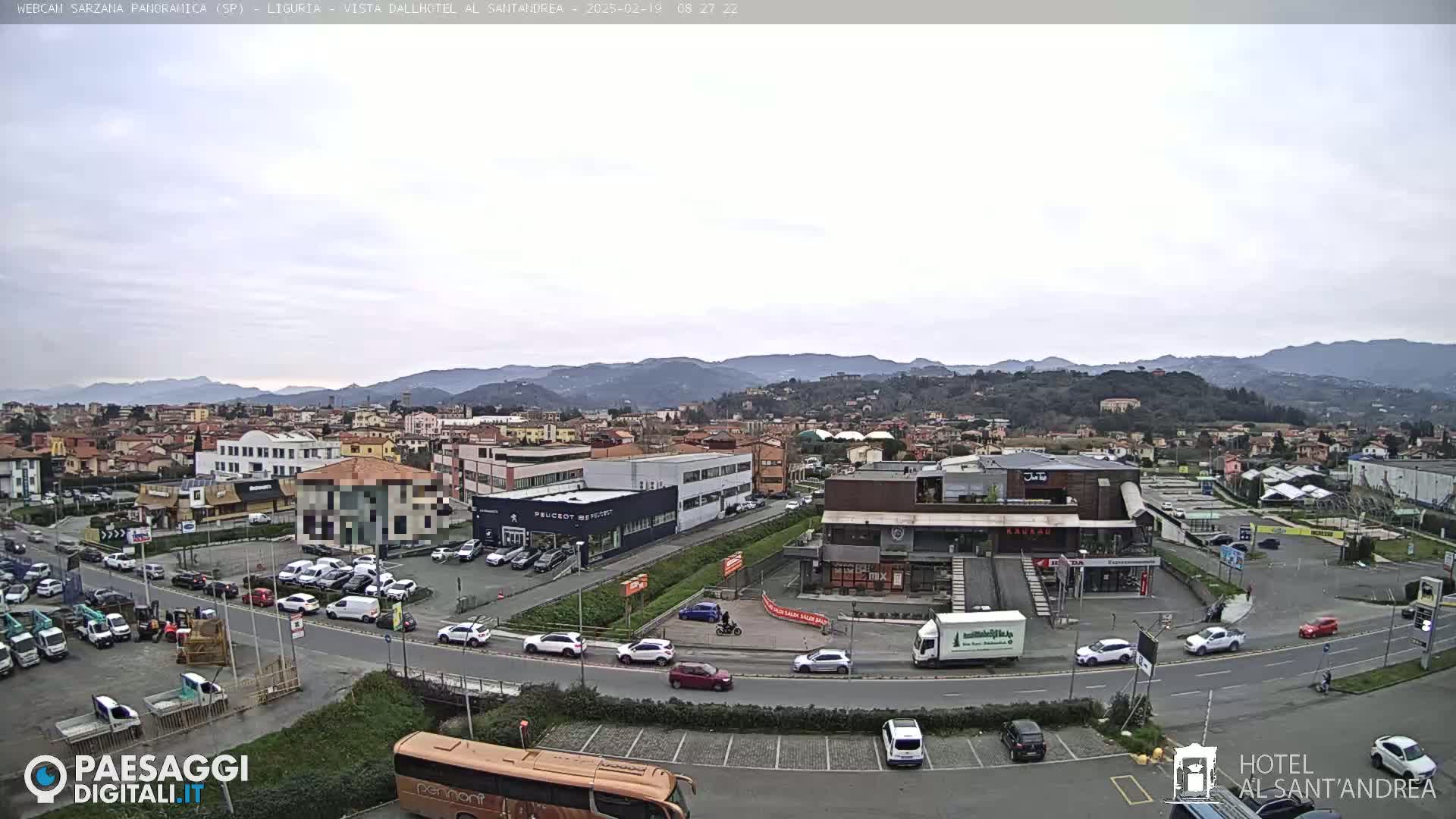 A partly cloudy day reveals a view of a town nestled in a valley, surrounded by distant mountains, with a road and parking lots filled with cars visible in the foreground.
