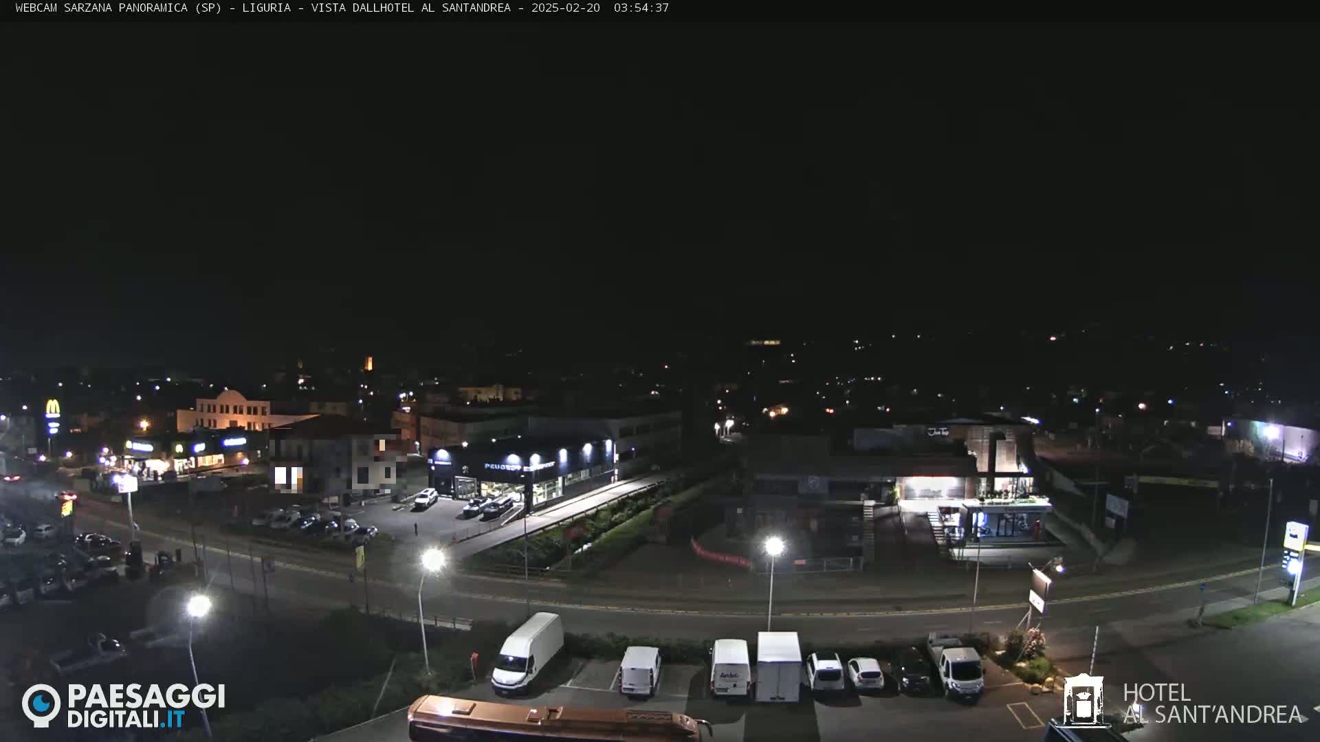 A nighttime aerial view of a town's outskirts showing roadways, parking lots with cars, and various lit buildings.