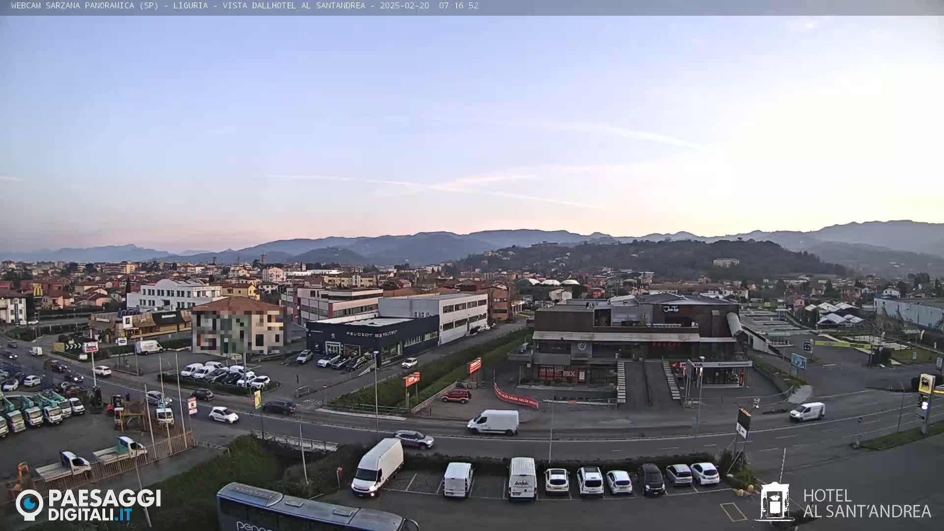 A panoramic view of a town nestled in a valley, under a clear, twilight sky, with numerous cars parked in lots and driving on roads.