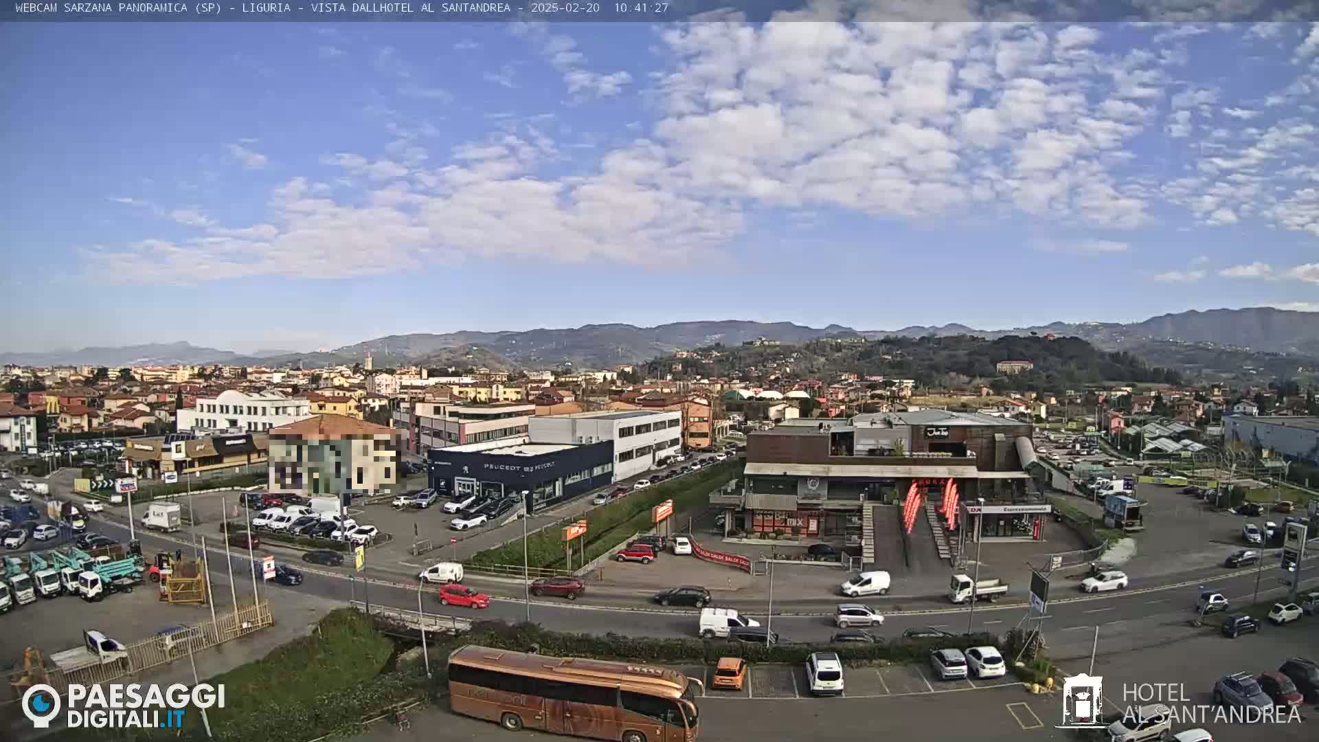 A partly cloudy day reveals a sprawling town nestled beside a mountain range, with numerous cars and a large bus visible in a parking lot near several buildings.