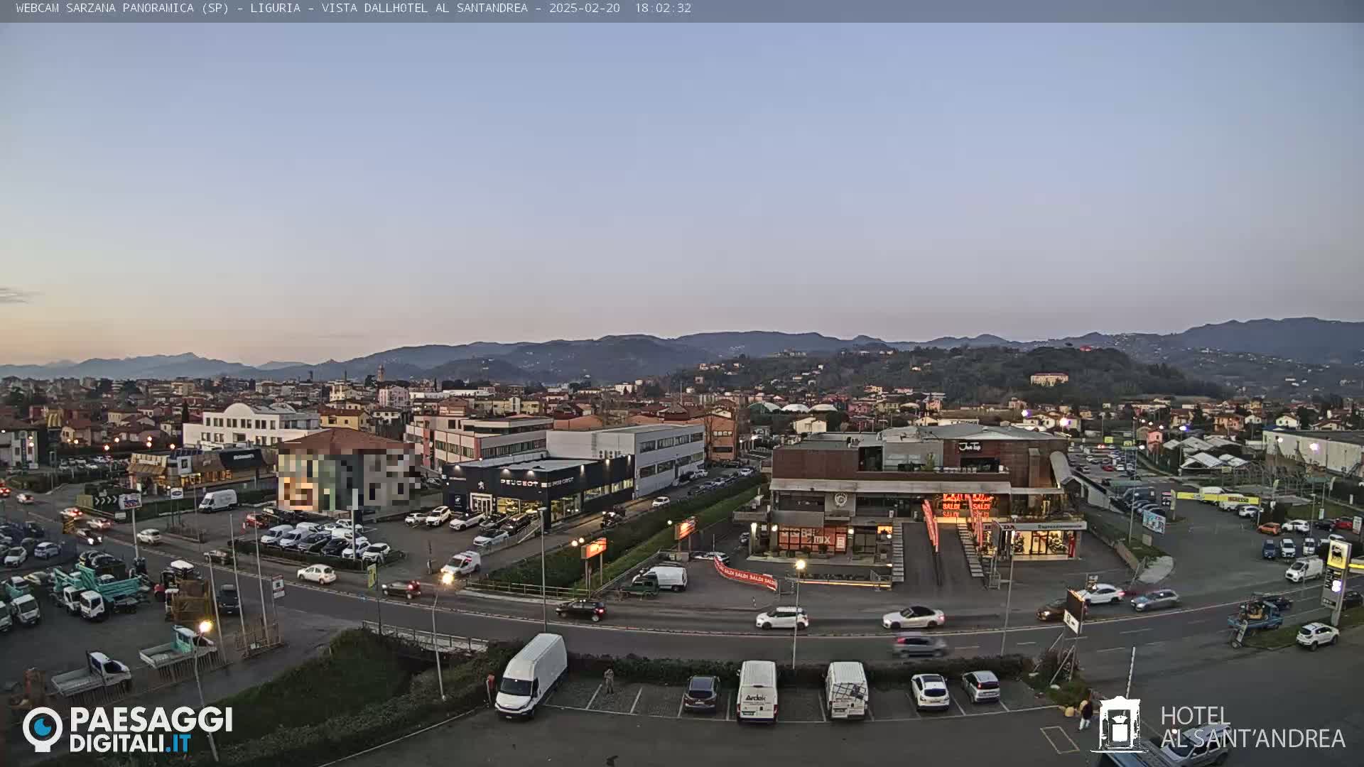 A twilight view of a town nestled in a valley, mountains in the background, with cars parked in lots surrounding various buildings.