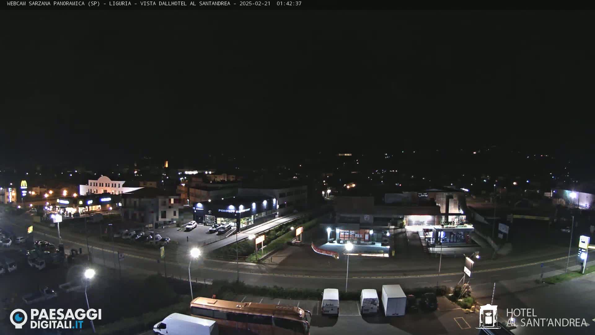 A nighttime aerial view of a town's outskirts, showing illuminated buildings, roads, and parked vehicles under a clear, dark sky.
