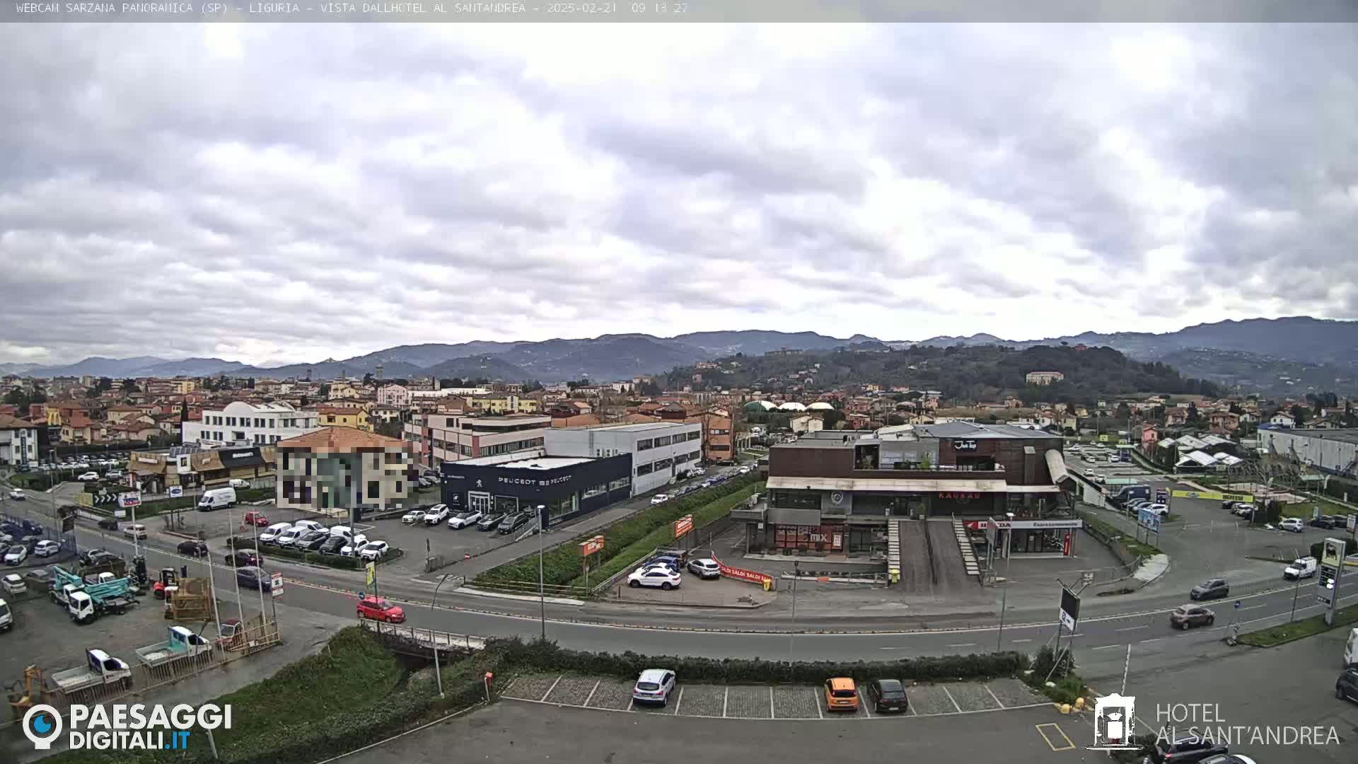 A mostly cloudy day reveals a sprawling town nestled in a valley, surrounded by rolling hills and mountains, with cars parked in lots near buildings and a road curving through the scene.