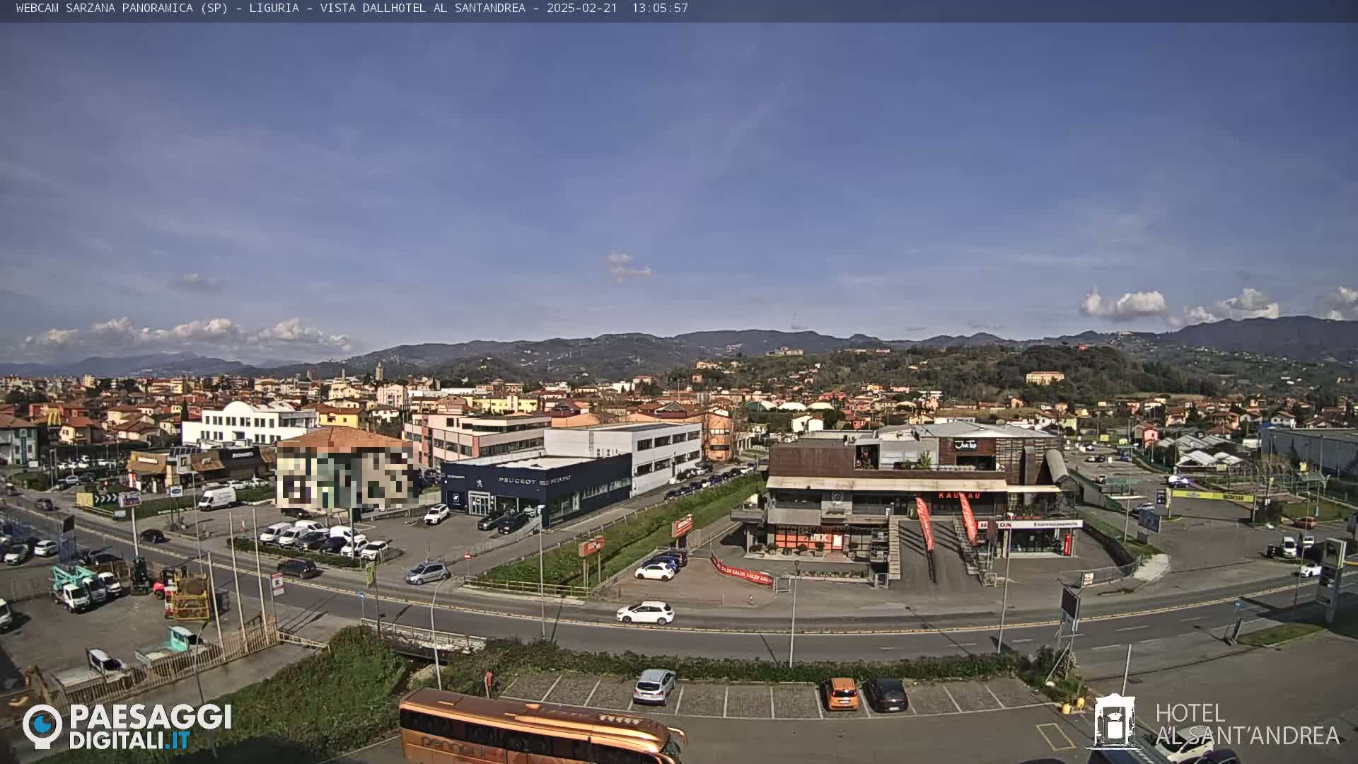 A sunny day reveals a panoramic view of a town nestled in a valley, with mountains in the background and various buildings including a car dealership and shops visible in the foreground.