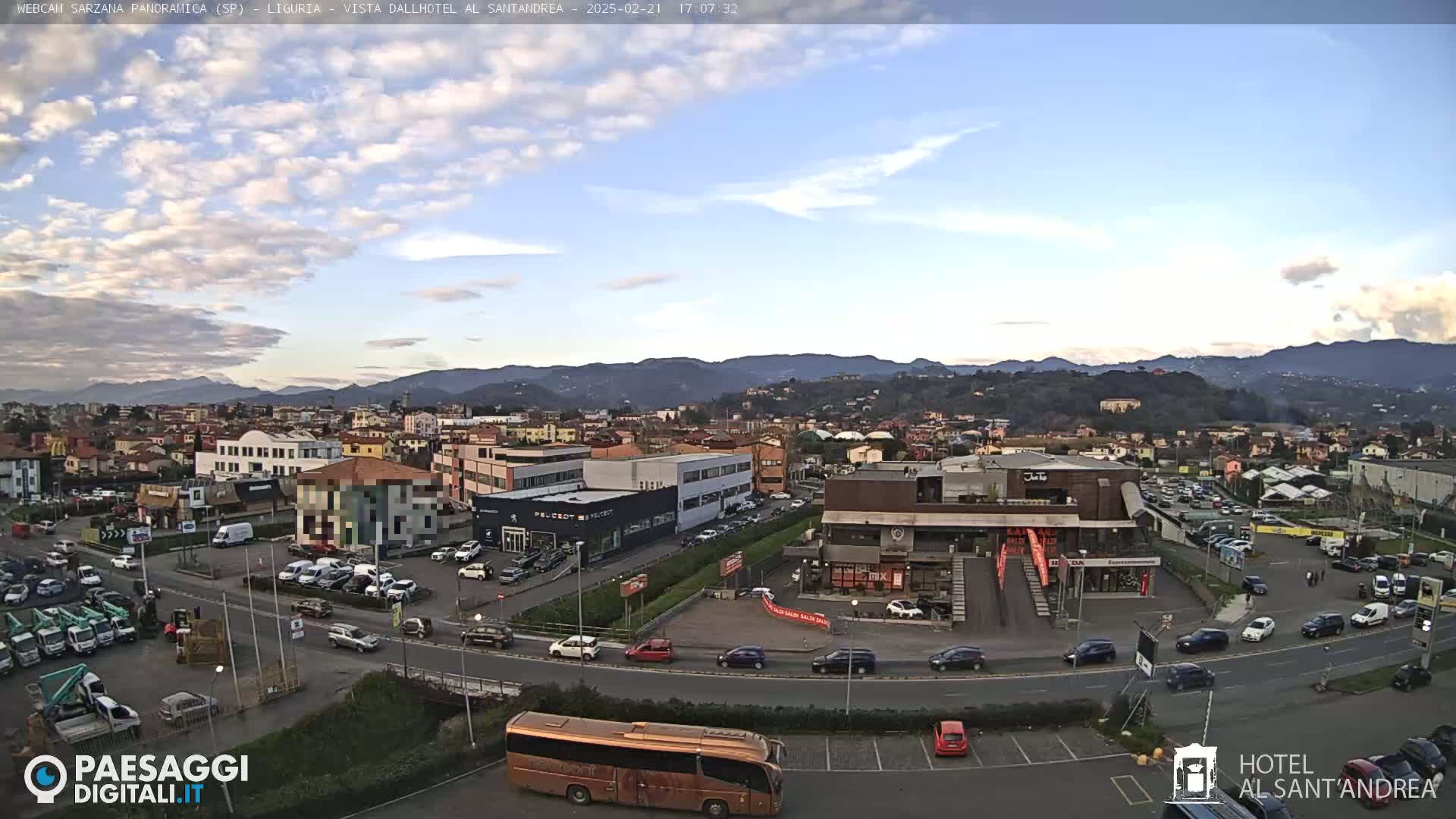 A partly cloudy daytime view of a town in a valley surrounded by mountains, with cars parked in lots and driving on roads.