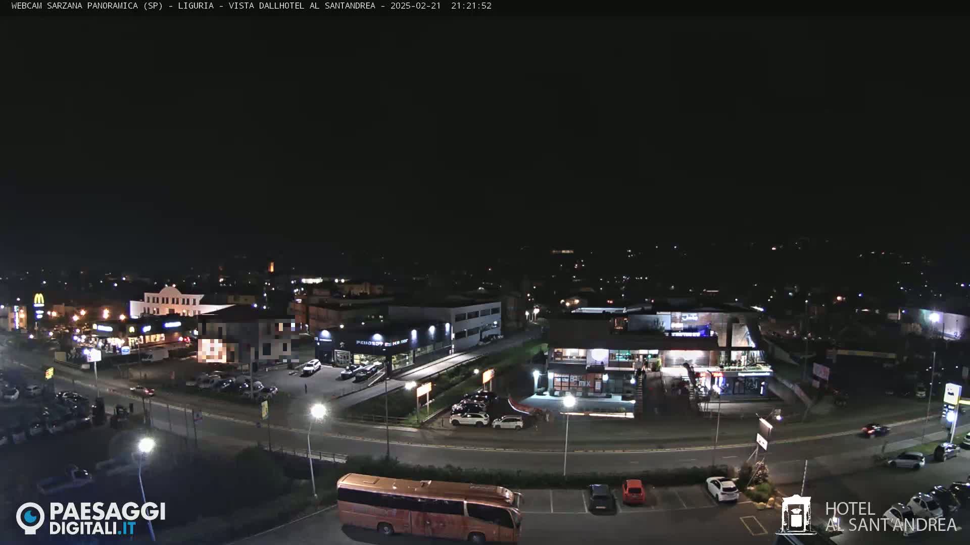 A nighttime, aerial view of a town's outskirts, showing illuminated buildings, roads, and parked vehicles.