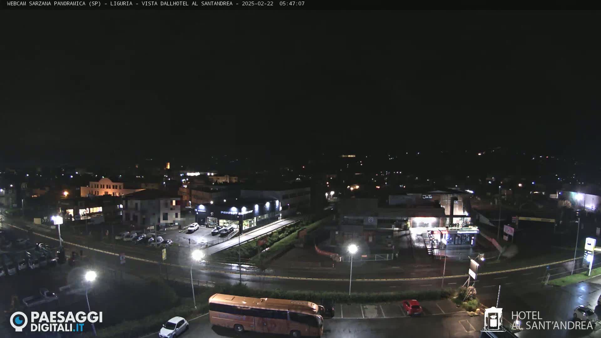 A nighttime, wet aerial view of a town with cars parked in lots, a large bus, and buildings illuminated by streetlights.
