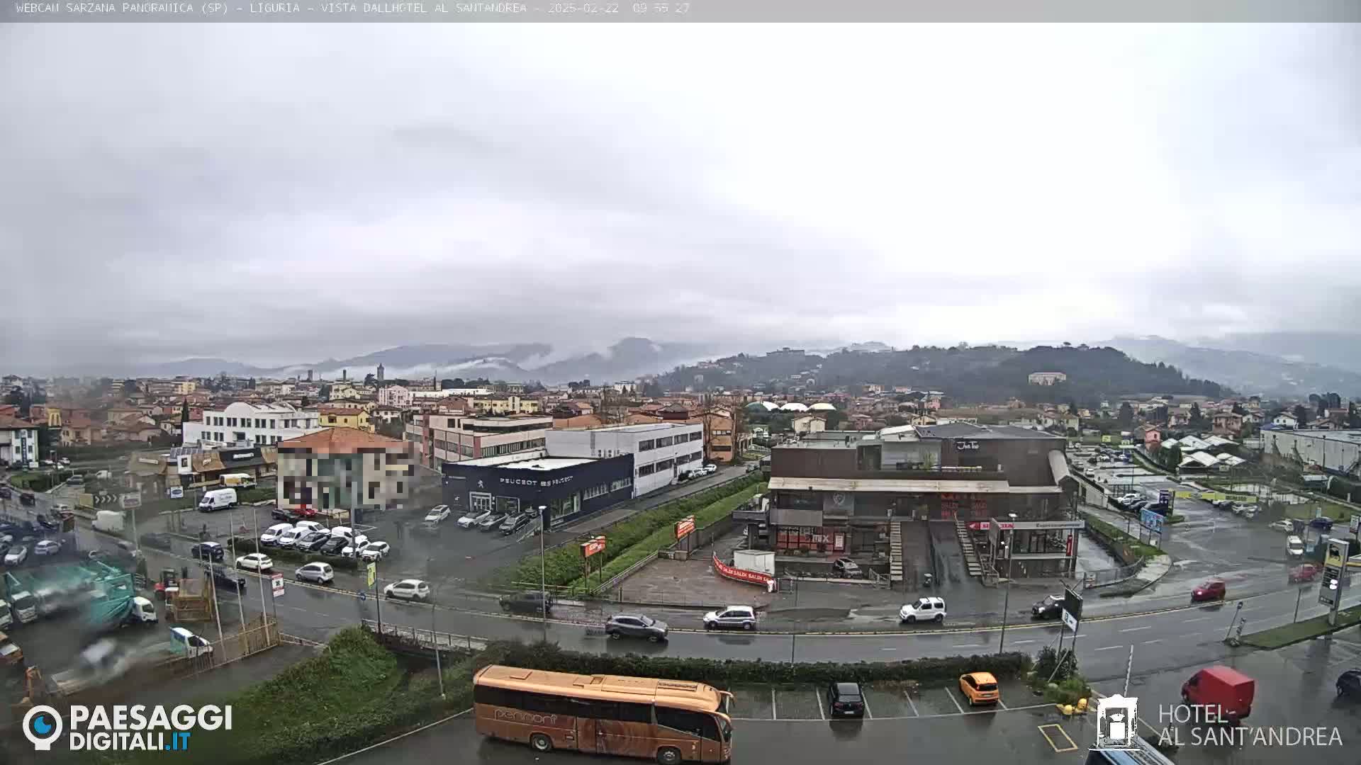 A wet, overcast city scene shows a wide road with cars and a bus, surrounded by buildings and parking lots, with hills in the background.