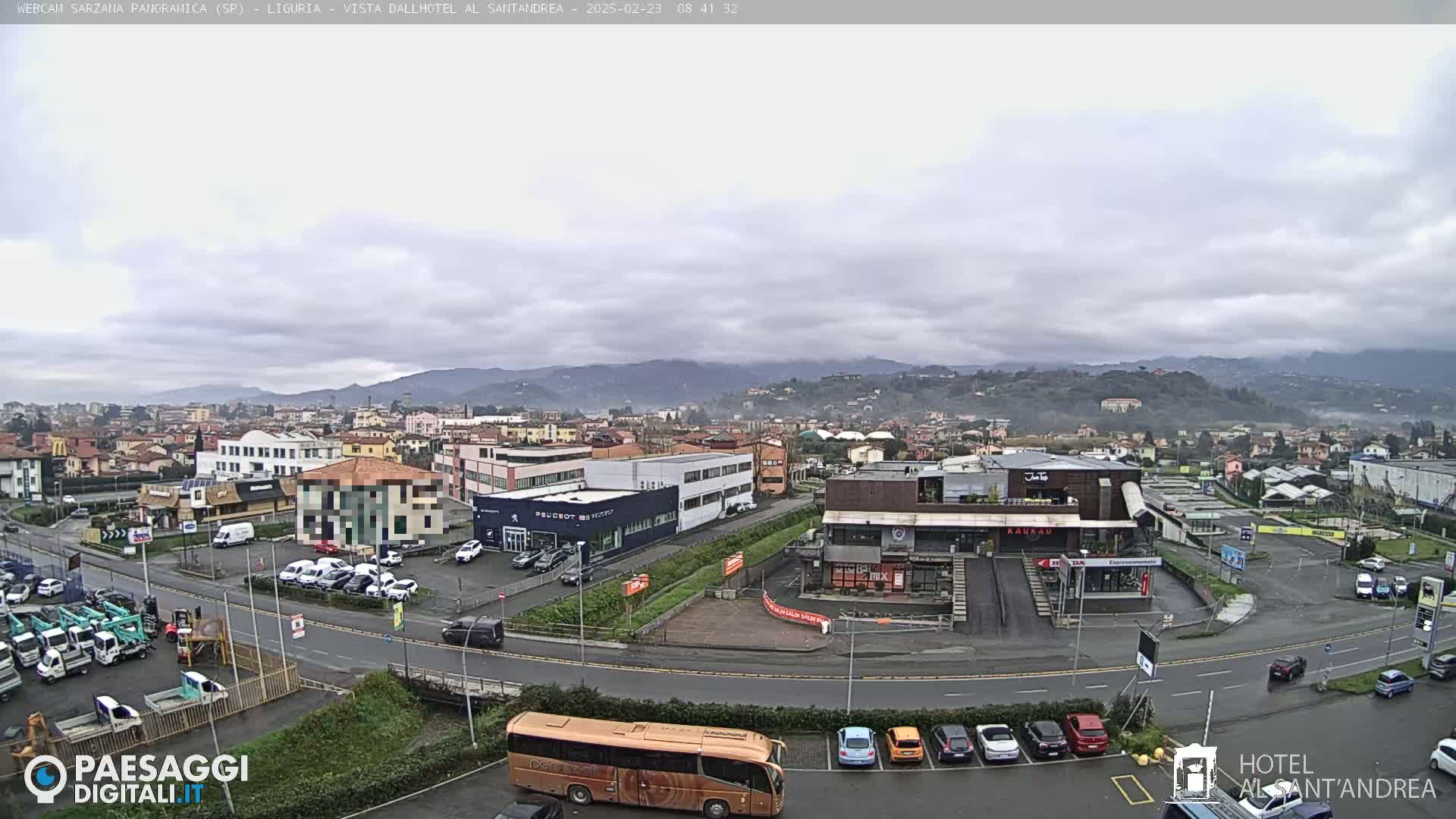 A panoramic view of a town on a cloudy day, showing buildings, roads, cars, and a bus, with hills in the background.