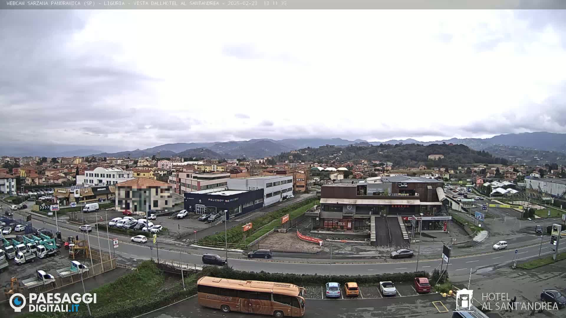A panoramic view of a town under a cloudy sky, showing buildings, roads, parked cars, and a bus, with hills in the background.