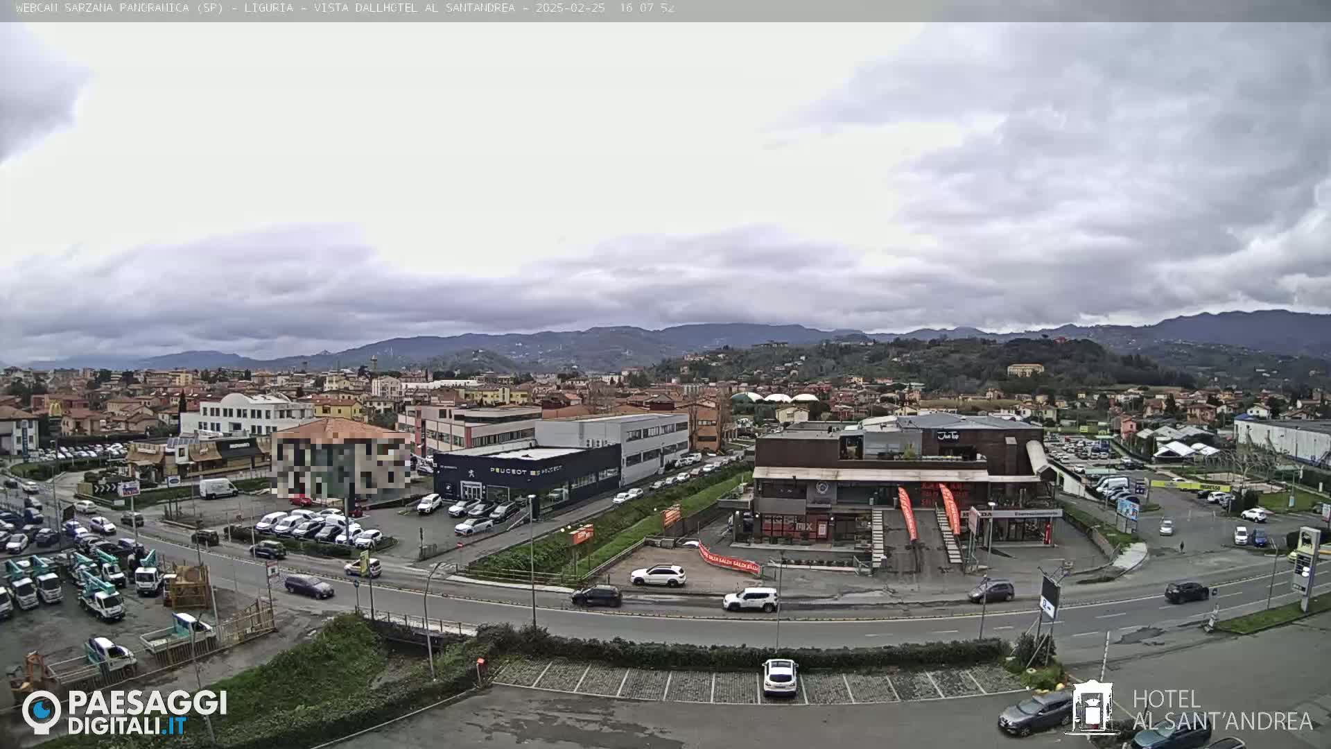 A wide shot of a town under a cloudy sky shows a cityscape with cars in parking lots and on roads, surrounded by hills in the distance.