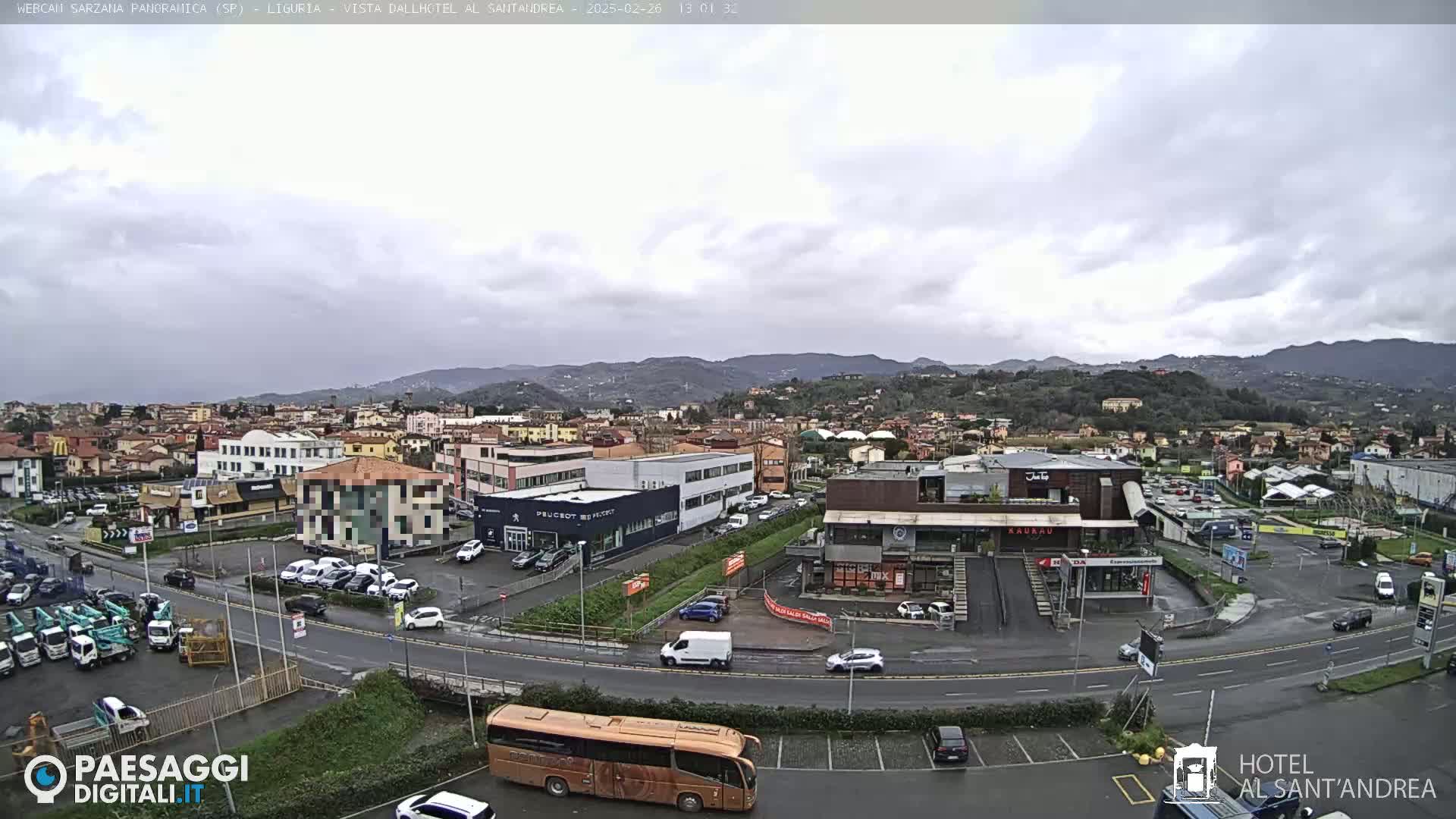 A long shot of a town under an overcast sky, showing a variety of buildings, vehicles, and a large bus in a parking lot.