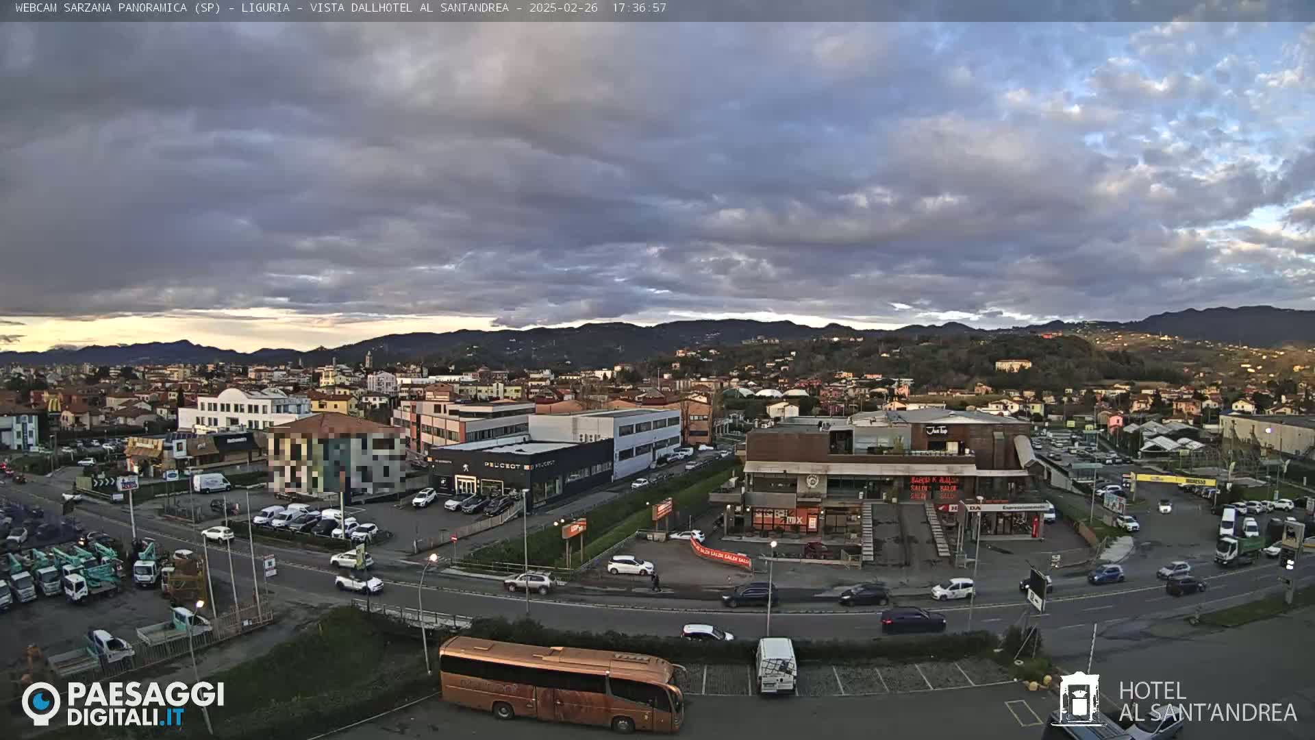 A partly cloudy evening view of a town situated in a valley, with mountains in the background and several cars in a parking lot and on roads.